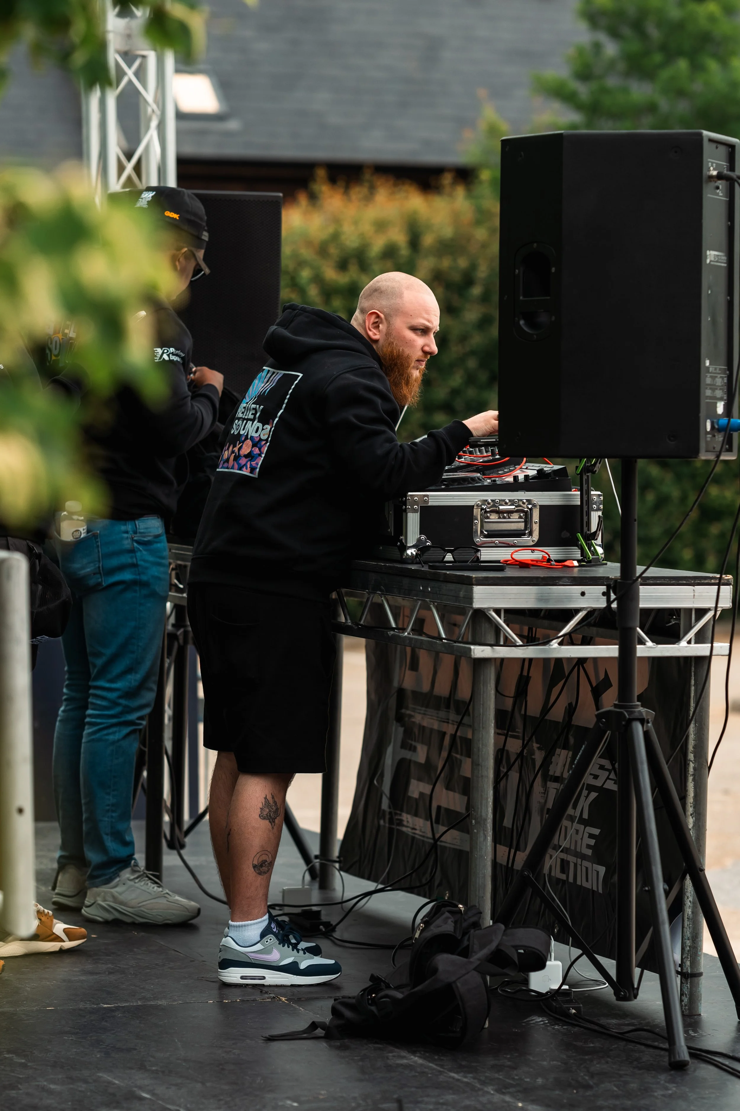 A man with a beard and tattoos on his leg working on DJ equipment at an outdoor event, with large speakers and other people nearby.