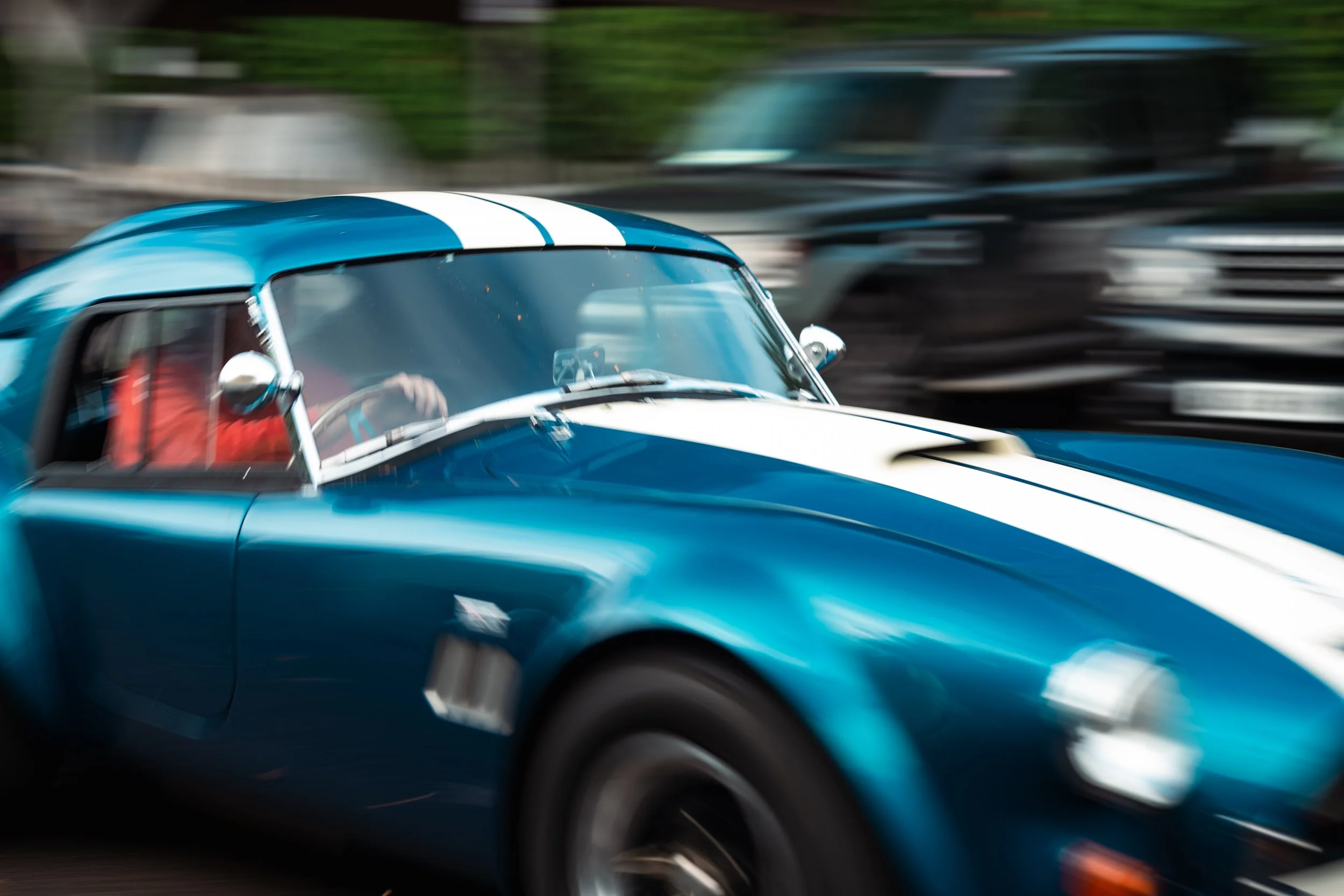 A blue vintage sports car with white racing stripes moving at high speed, with blurred background and wheels indicating motion.