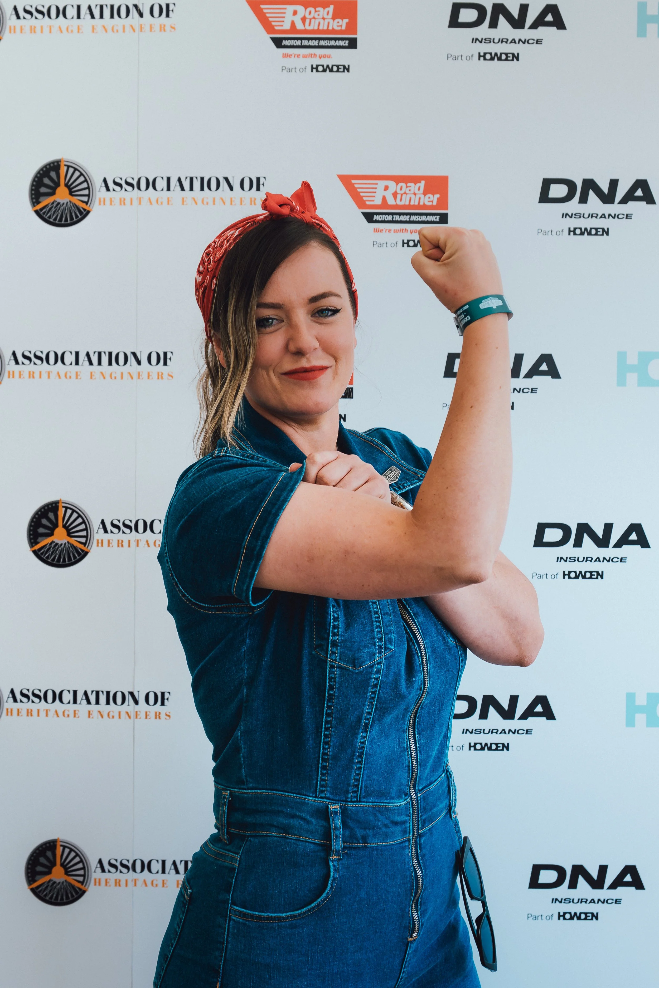 A woman with a red bandana and denim shirt flexing her right arm to show her bicep, standing in front of a backdrop with logos including Association of Heritage Engineers, Road Runner, DNA Insurance, and Holden.