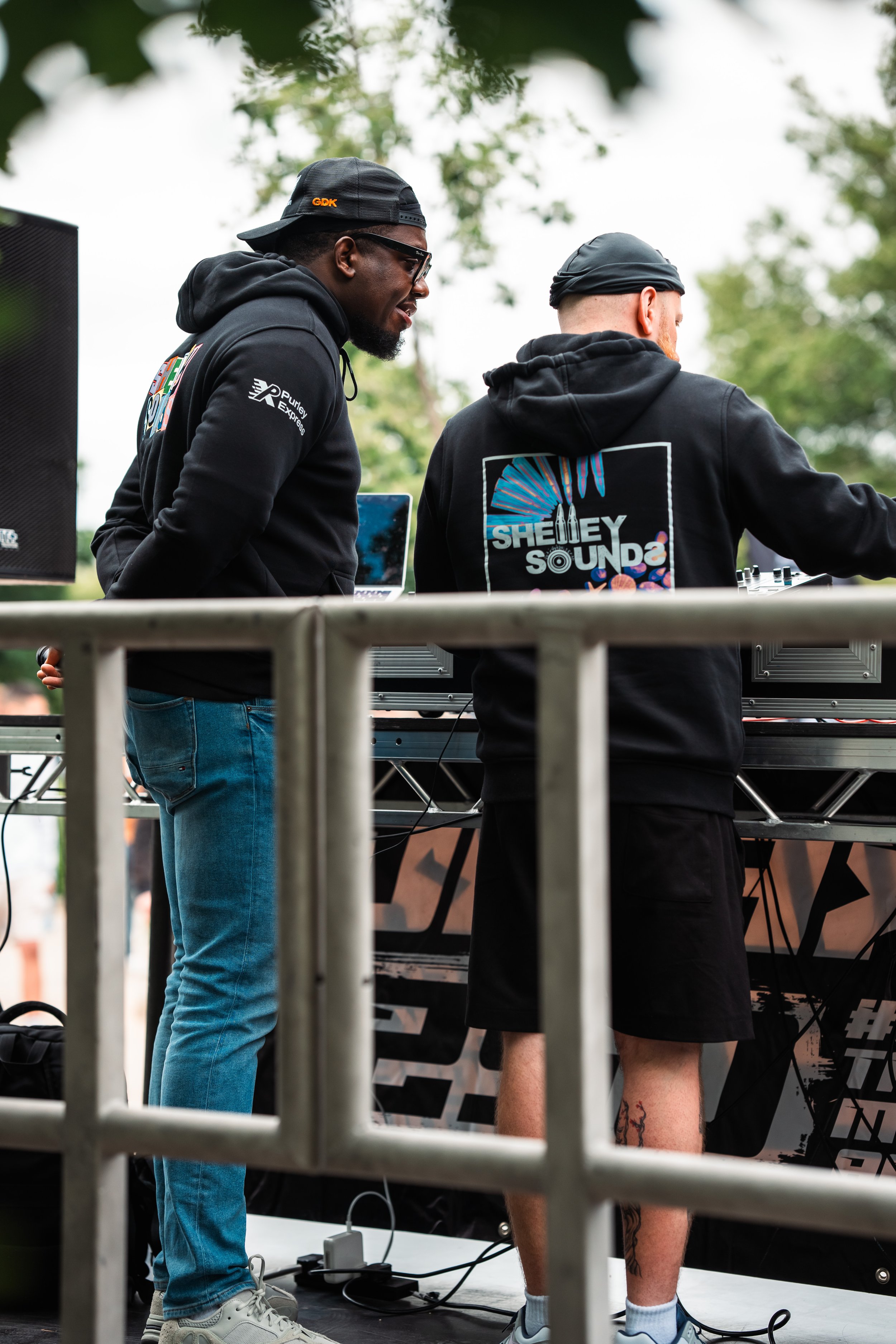 Two men standing at a DJ booth outdoors, surrounded by trees. One is wearing a black hoodie and blue jeans, the other in a black hoodie with a graphic on the back, black shorts, and a black head covering. They appear to be operating DJ equipment.