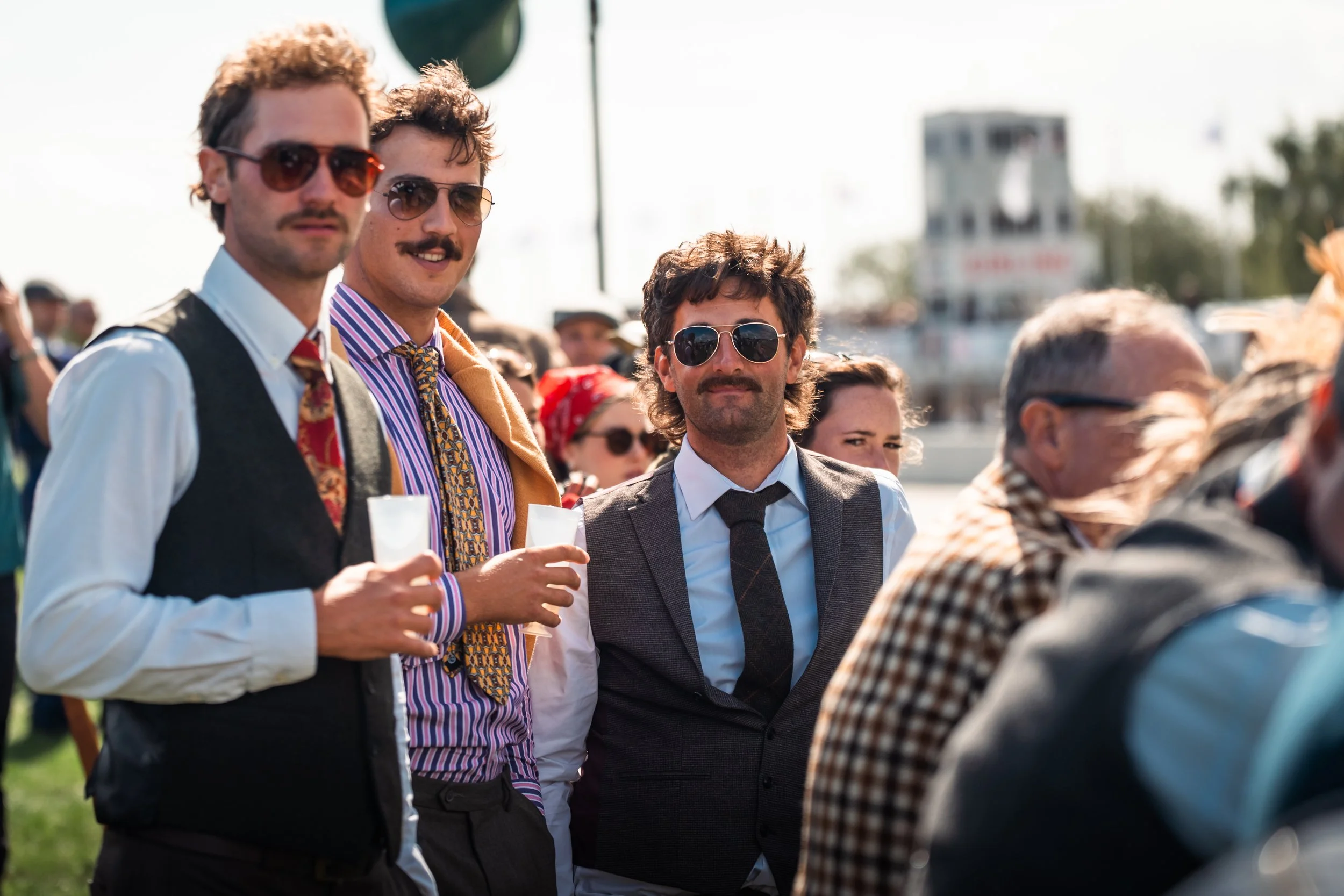 Three men with sunglasses and mustaches standing outdoors at an event, surrounded by a crowd, with a blurred green background and some structures in the distance.