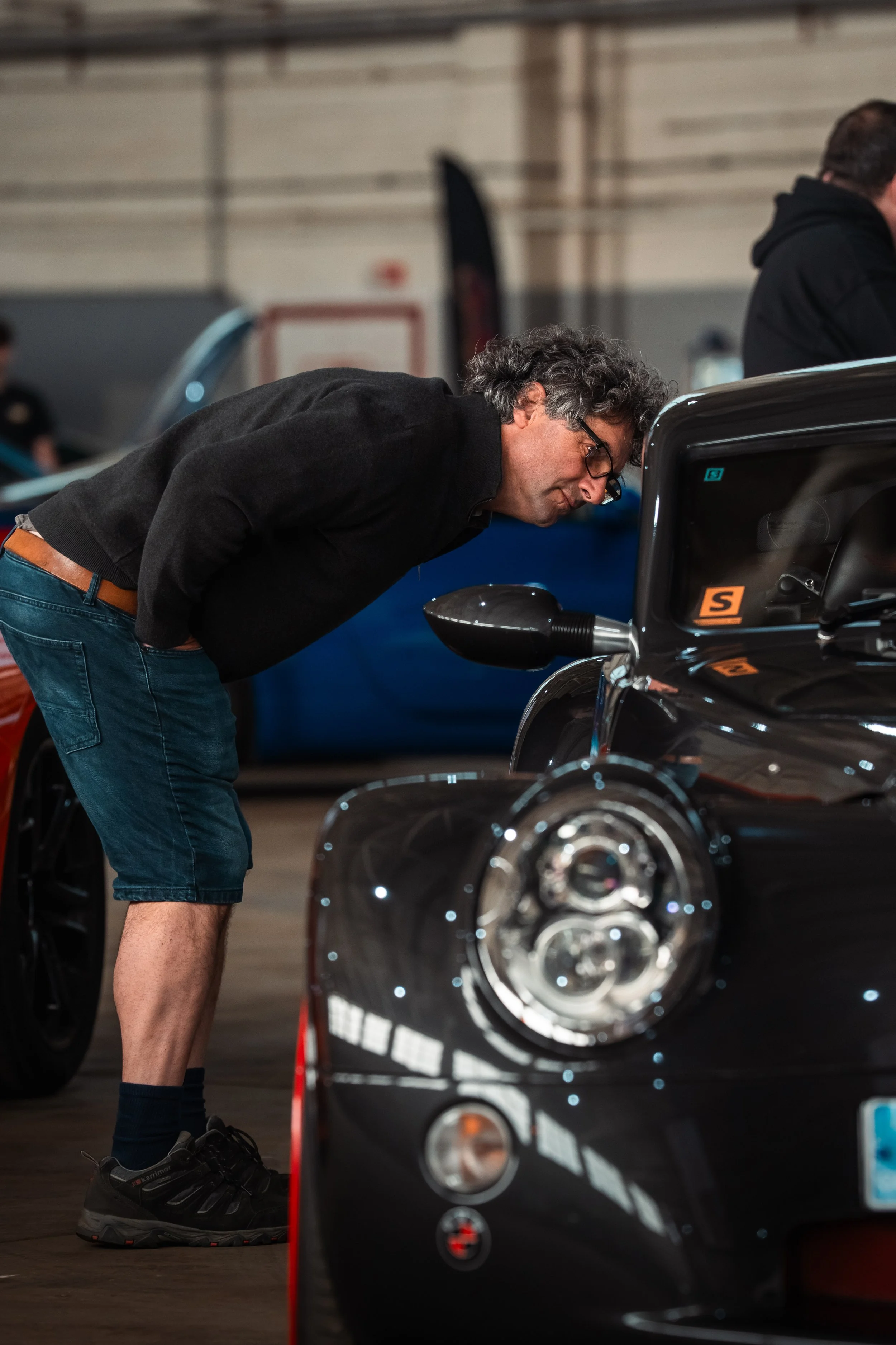 A man with curly gray hair, glasses, wearing a black long-sleeve shirt, blue shorts, and black sneakers, leaning forward and closely inspecting a black motorcycle inside a well-lit indoor space.