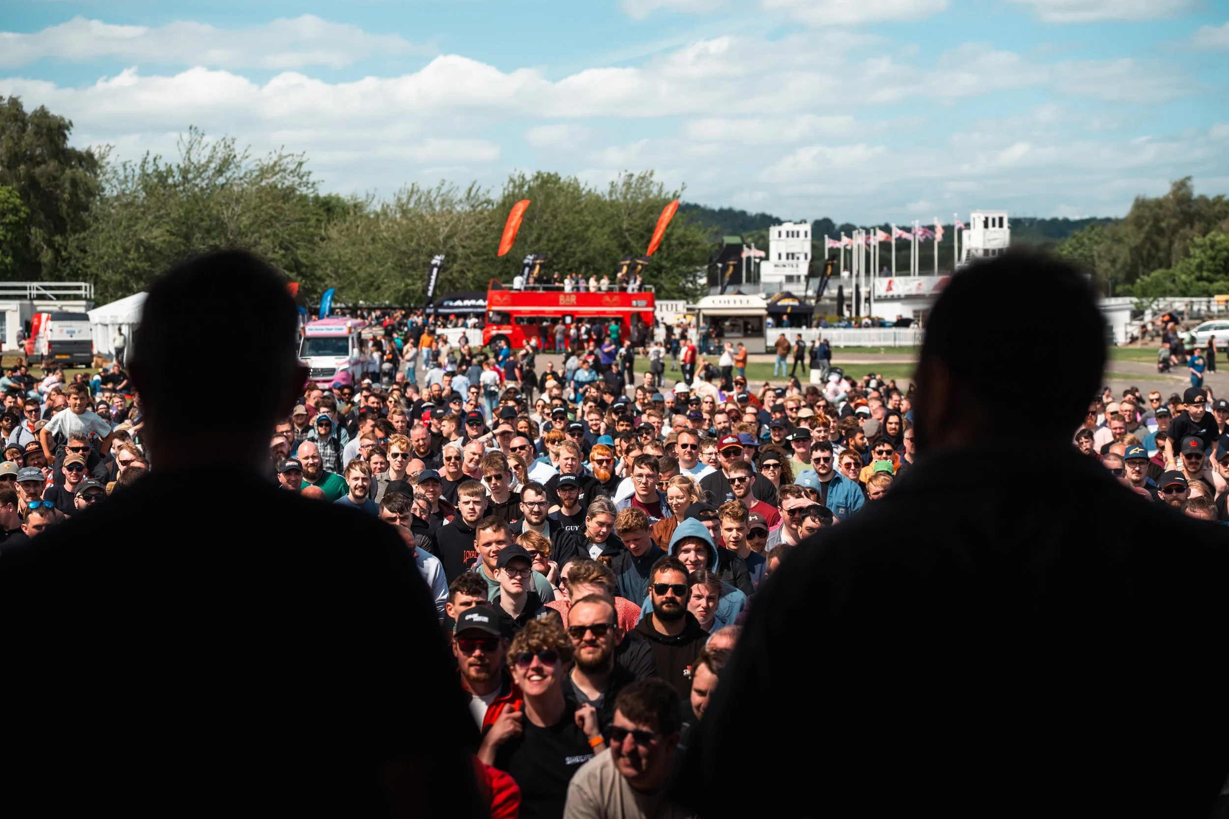 A large crowd of people gathered outdoors at an event with flags and banners, with some structures and trees in the background, seen from behind two silhouetted heads.