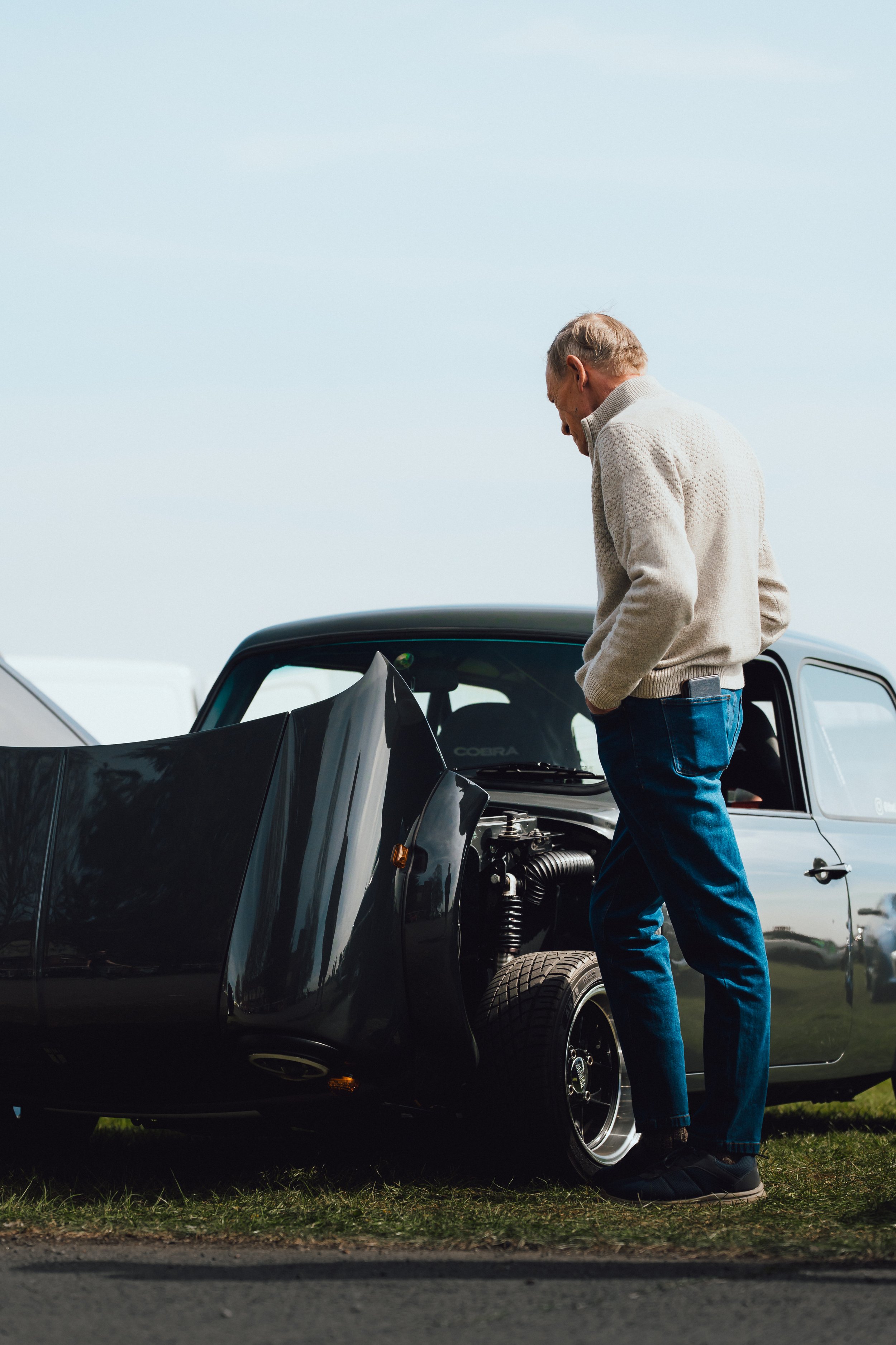 An elderly man with gray hair wearing a light-colored sweater and jeans is looking under the hood of a black sports car parked outdoors on a grassy area.