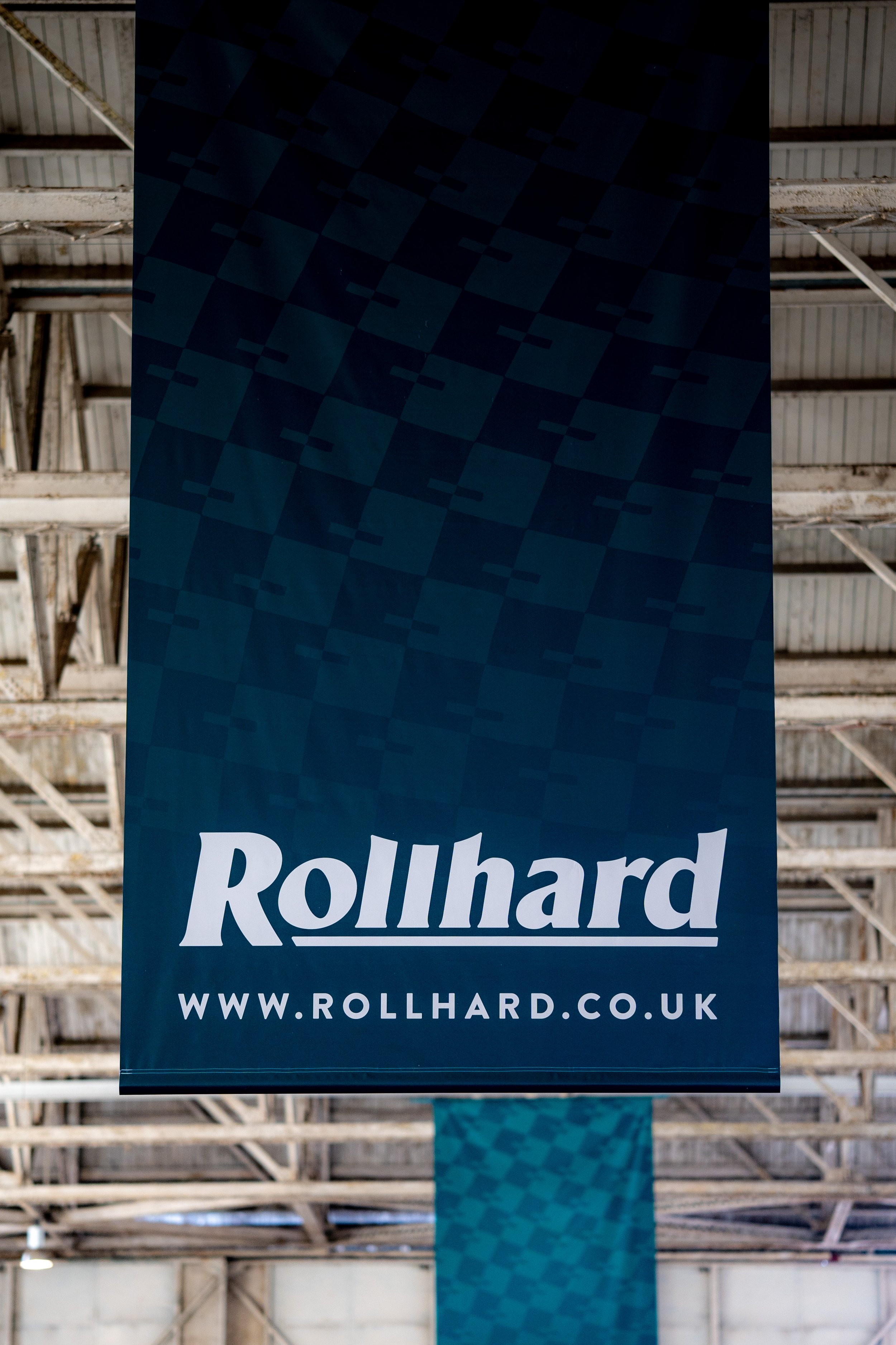 Blue vertical banner with the word 'Rollhard' in white text and the website URL 'www.rollhard.co.uk' at the bottom, hanging inside a warehouse with a wooden roof structure.
