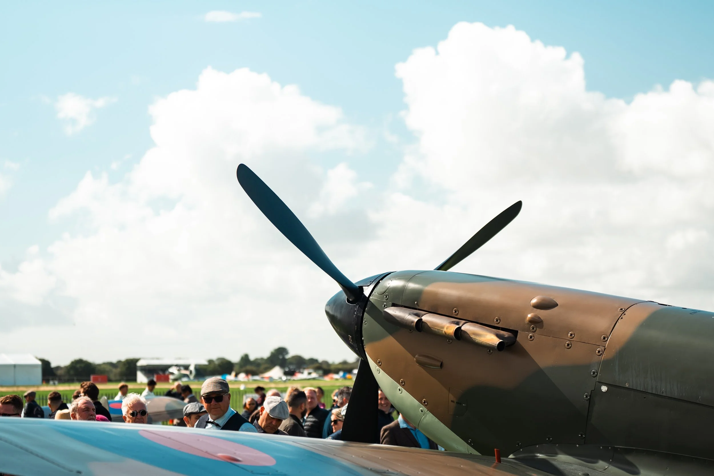 People gathered at an airshow observing a vintage military aircraft with a propeller. The sky is partly cloudy.