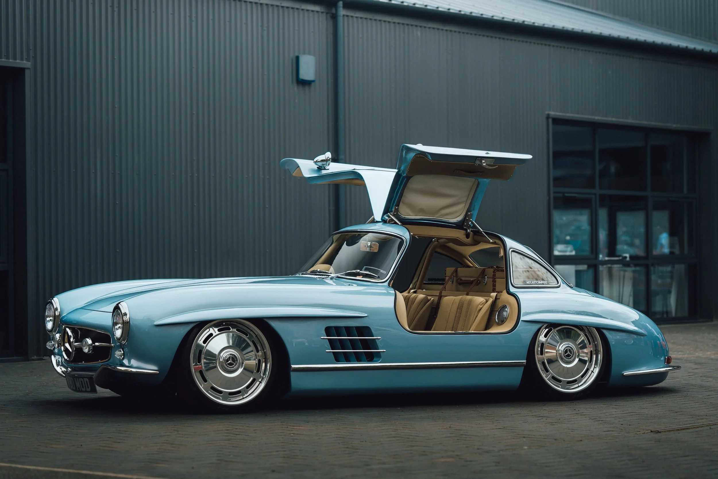 A vintage light blue convertible car with its trunk open, parked outside a modern industrial building with corrugated metal walls.