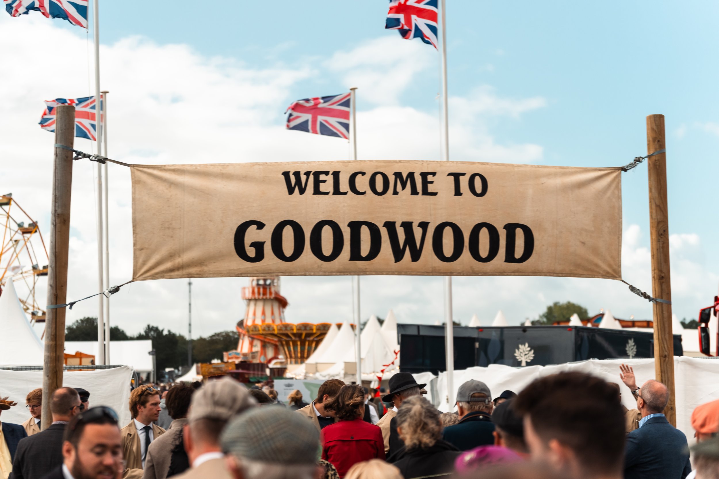 Crowd of people at an outdoor event with a banner that says 'Welcome to Goodwood' and Union Jack flags flying above.