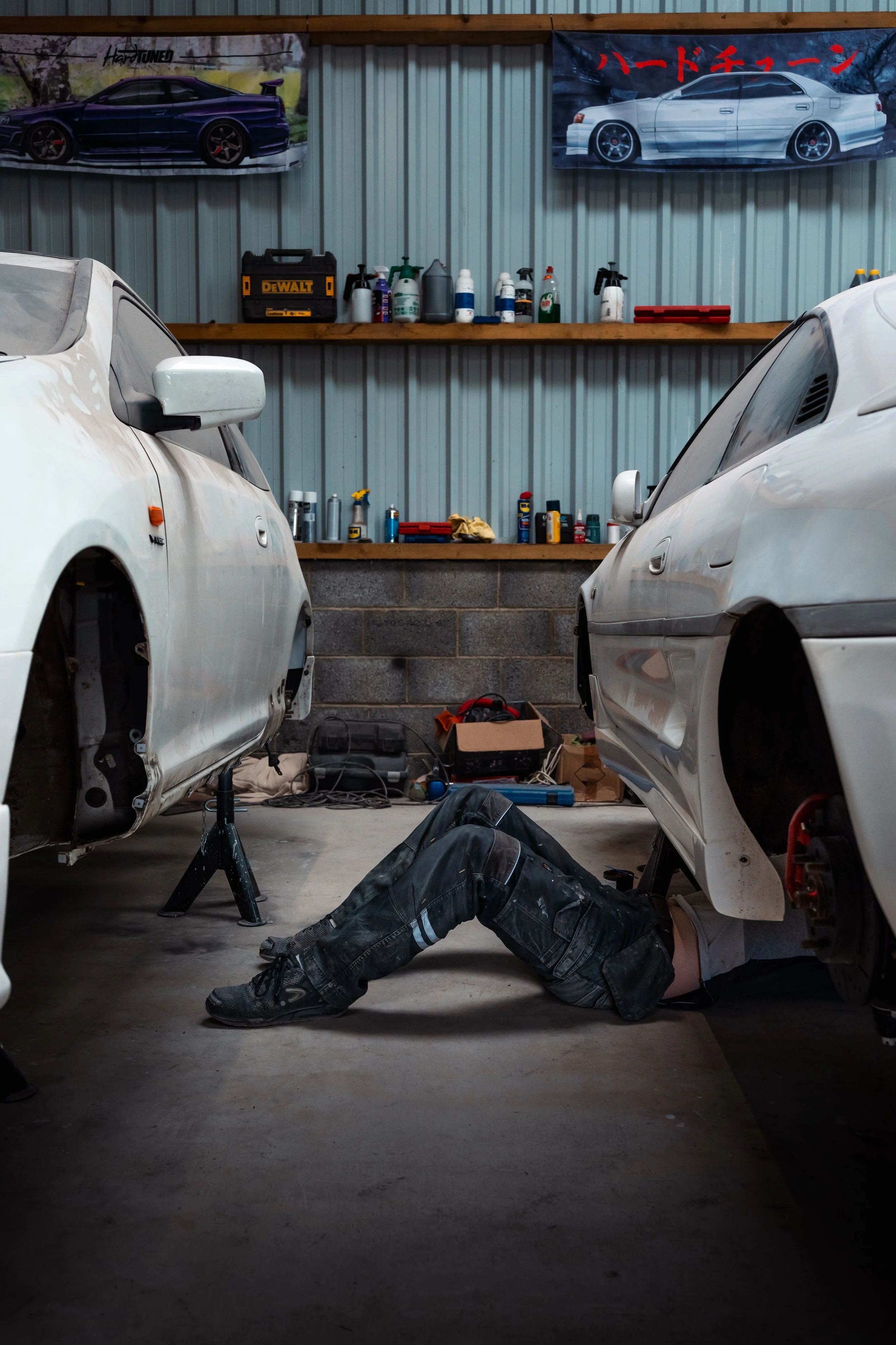A man working underneath two white cars in a garage, surrounded by tools and cleaning supplies on shelves.