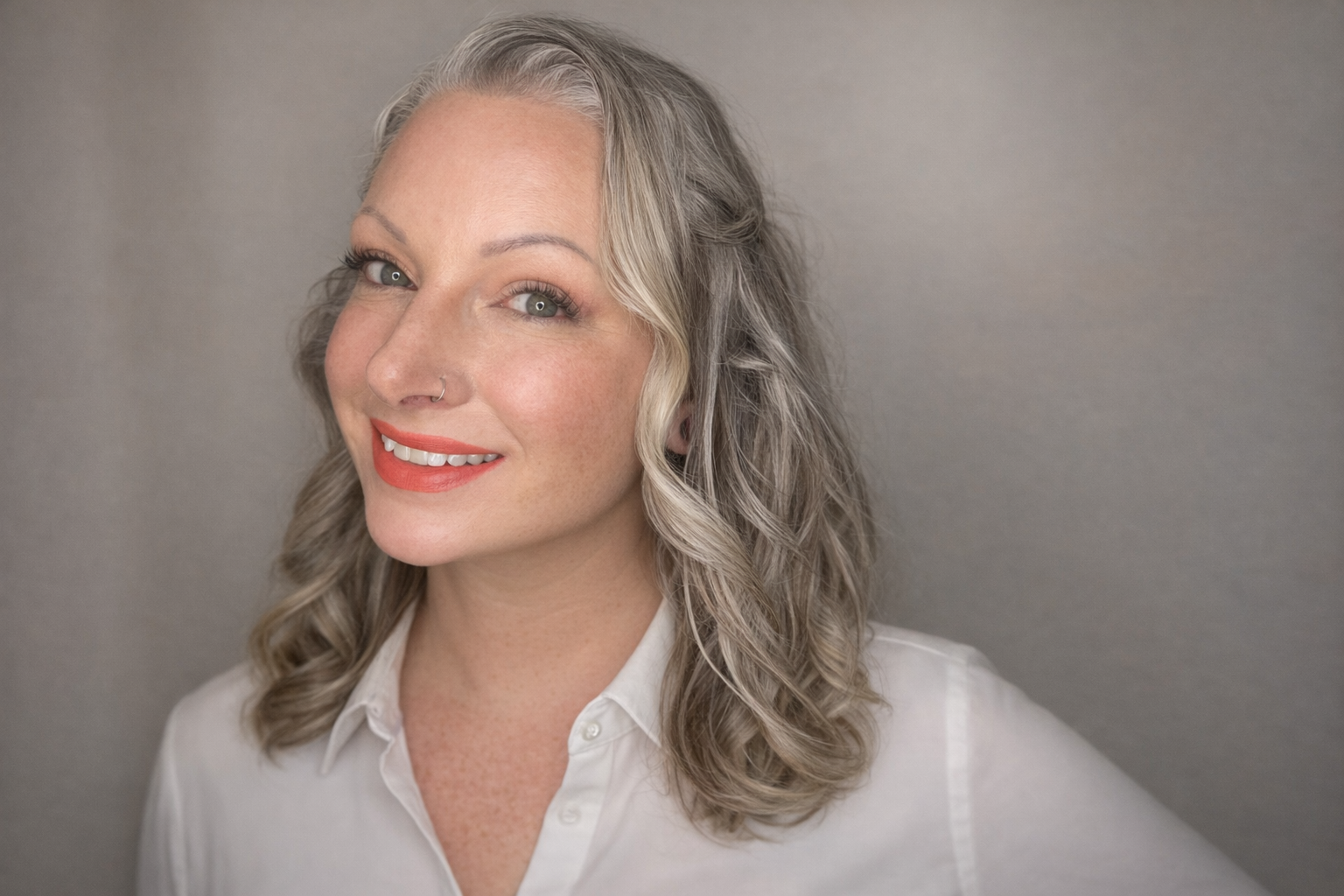 A woman with grey wavy hair, wearing a white collared shirt, smiling and looking slightly to the left, with a light grey background.