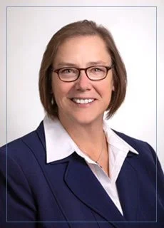 Professional portrait of a woman with short brown hair, glasses, wearing a navy blazer over a white collared shirt, smiling against a plain white background.