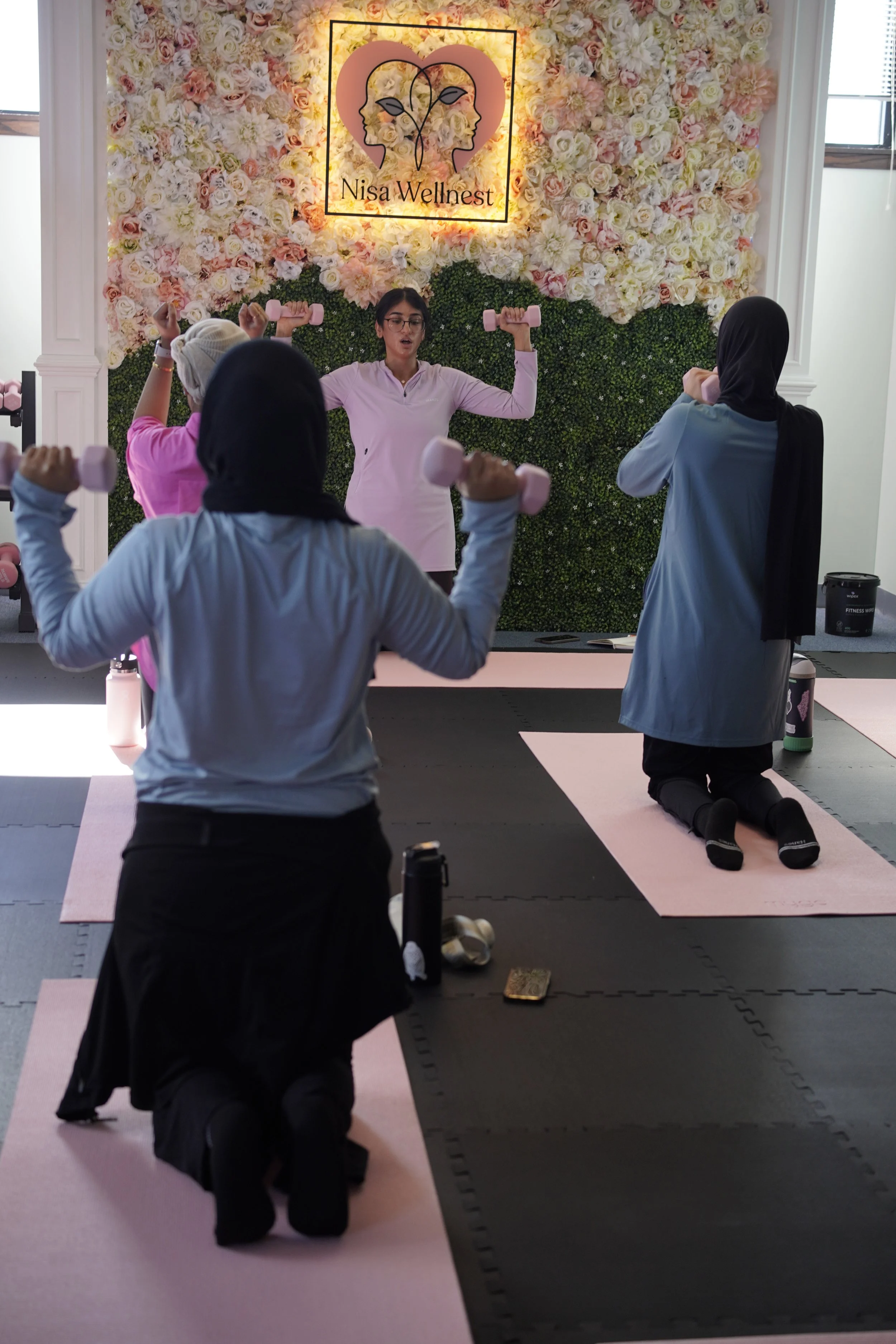 A fitness instructor leading a workout class with students holding pink dumbbells in a room decorated with a flower wall and a sign reading 'Nisa Wellness'.