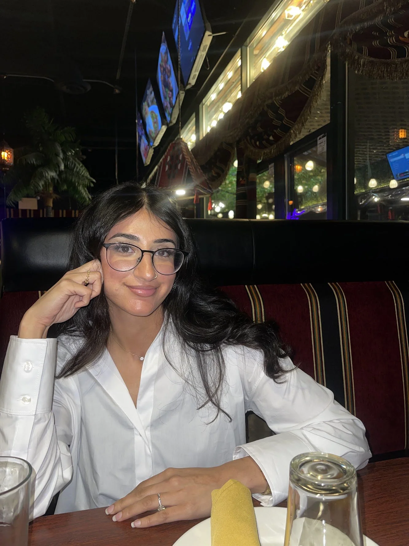 A woman with glasses and long dark hair is sitting at a restaurant table in a white blouse, smiling at the camera. The restaurant interior has dim lighting, colorful curtains, and TV screens on the wall.