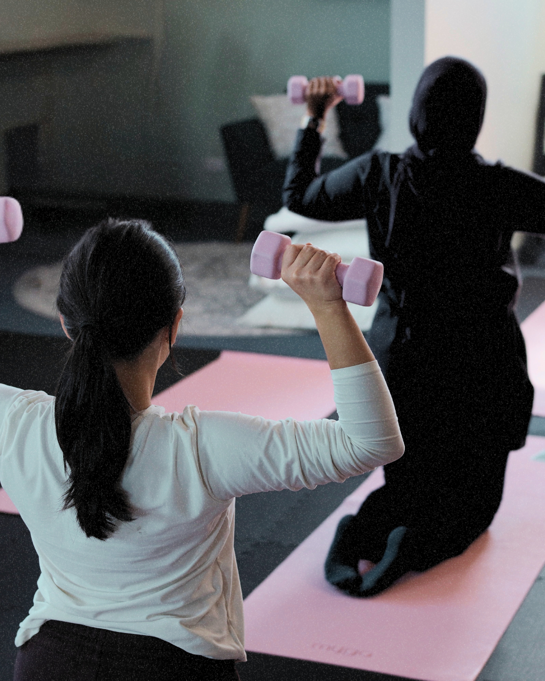 A woman and a man exercising with pink dumbbells in a home gym, with a yoga mat and a mirror in the background.