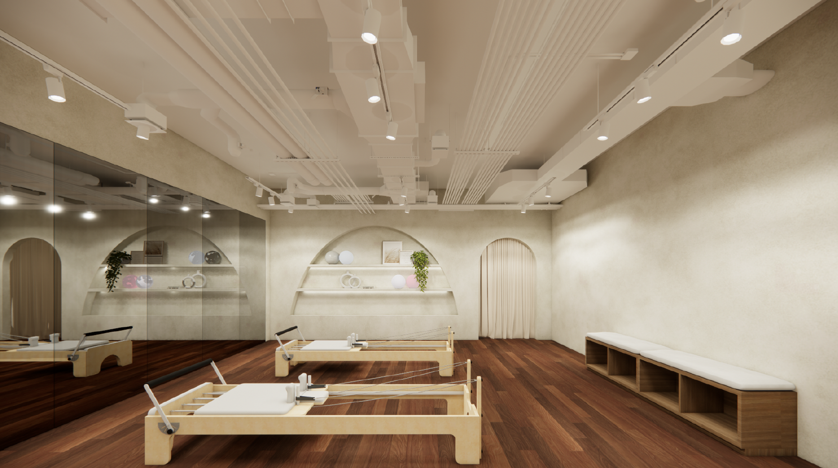 A dance or Pilates studio with wooden floors, light-colored walls, a large mirror on the left wall, and two reformer Pilates machines in the center. The ceiling has exposed white pipes and spotlights, and there are built-in benches with cushions on t