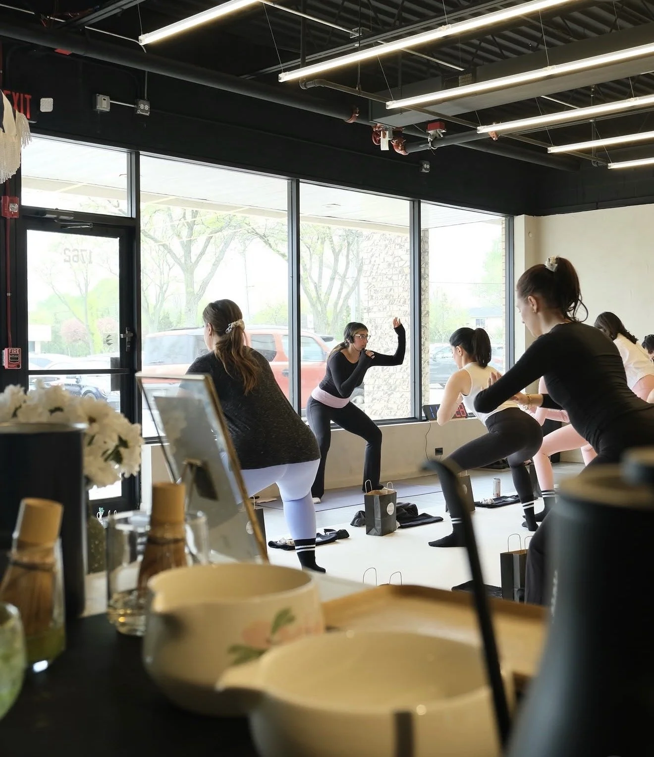 Women participating in a fitness class in a studio with large windows, led by an instructor demonstrating arm exercises.