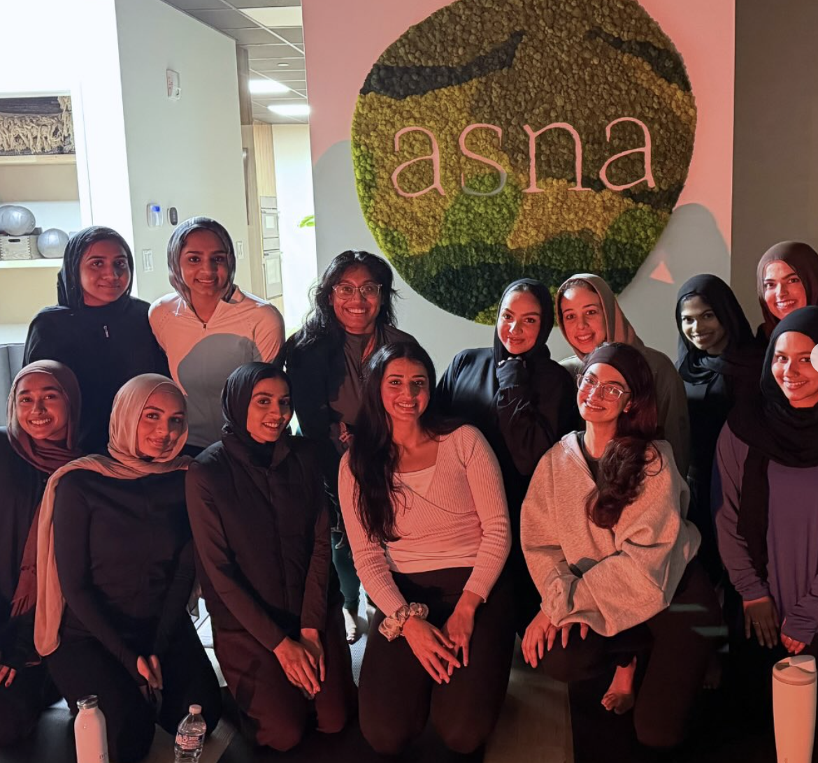 Group of women posing in front of a wall with a circular floral decoration and the word 'asna'.