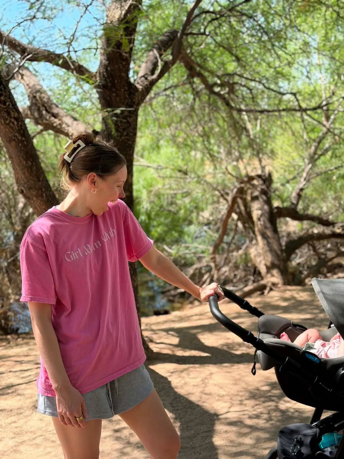 A female Naturopathic Doctor in a pink t-shirt and gray shorts pushing a baby stroller on a trail in a wooded park with green trees and a dirt path.