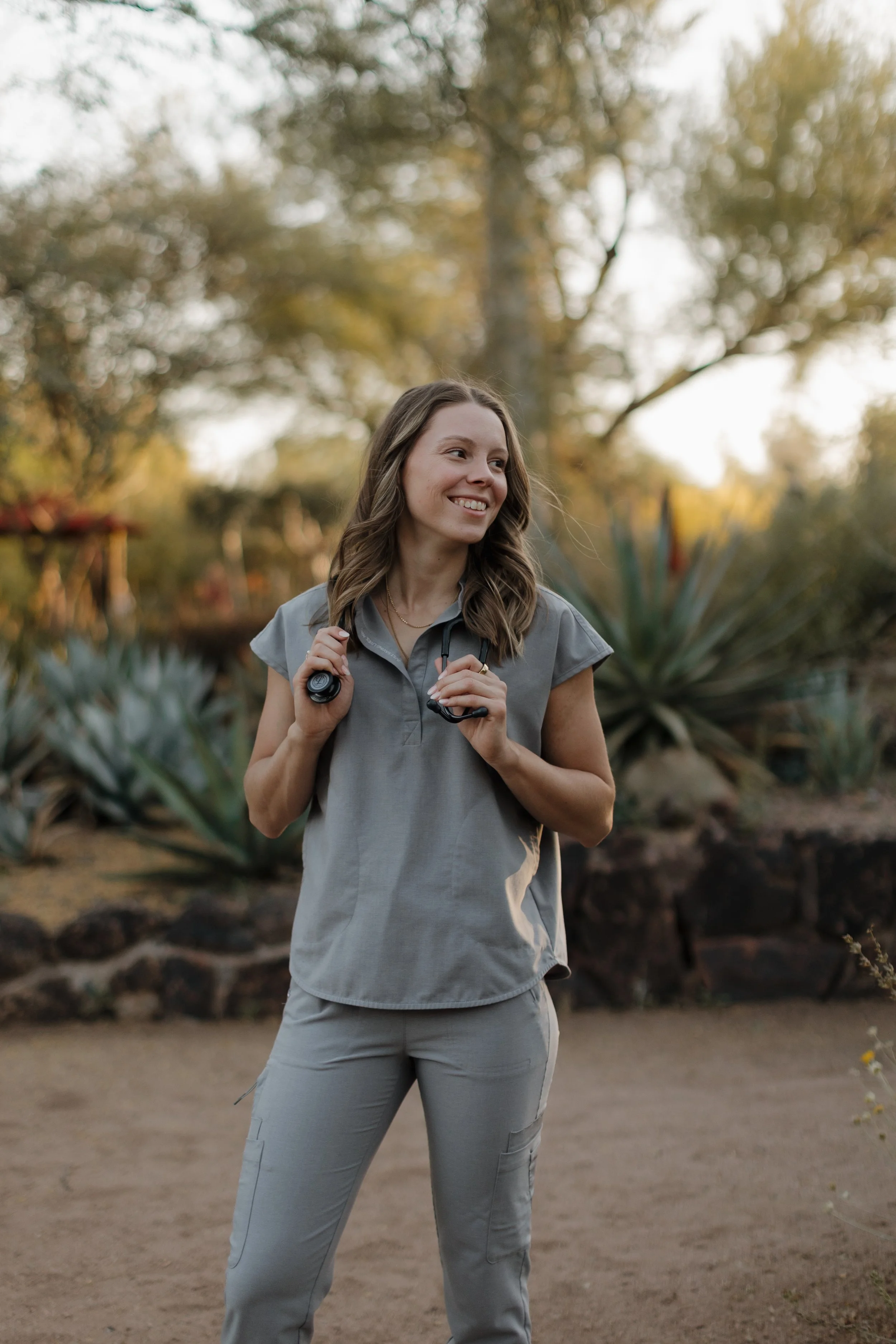 A female naturopathic doctor dressed in a light gray work uniform, smiling, holding a stethoscope, standing outdoors with desert plants and trees in the background.