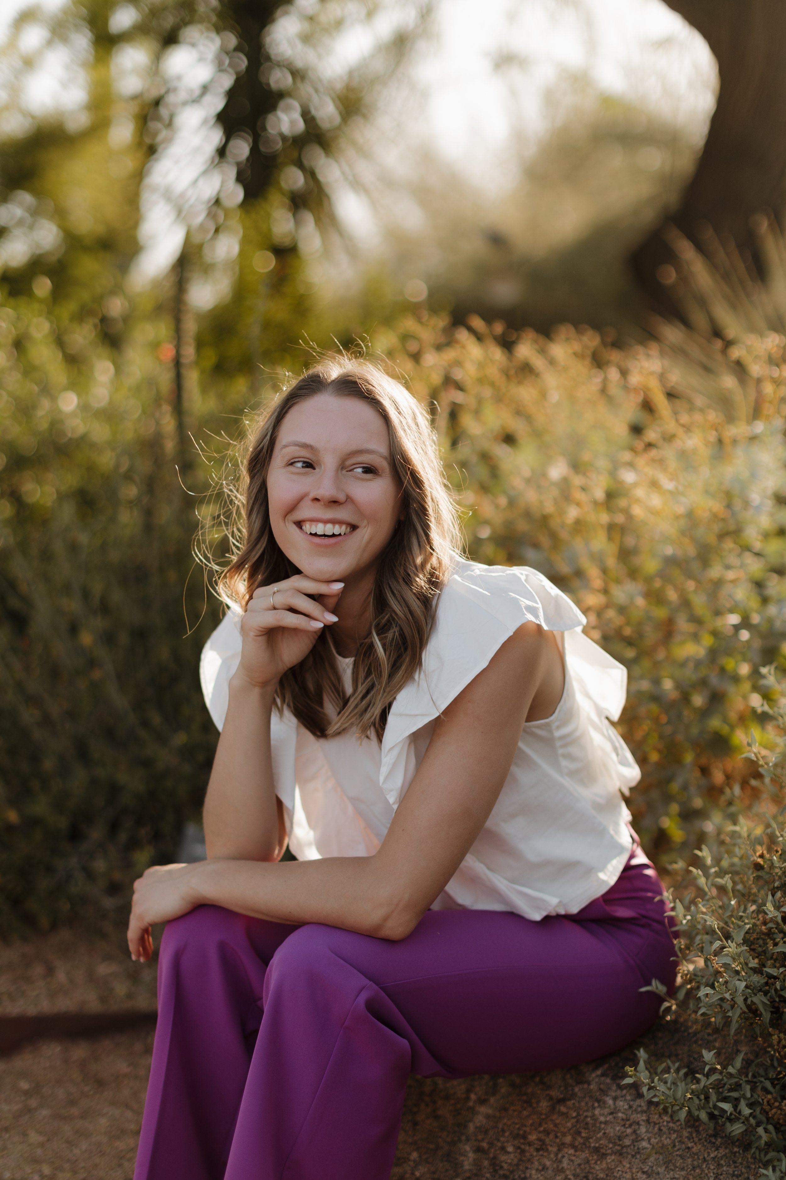 A Naturopathic Doctor, woman, with long wavy hair, smiling, sitting outdoors in a natural setting with trees and bushes in the background, wearing a white blouse and purple pants.