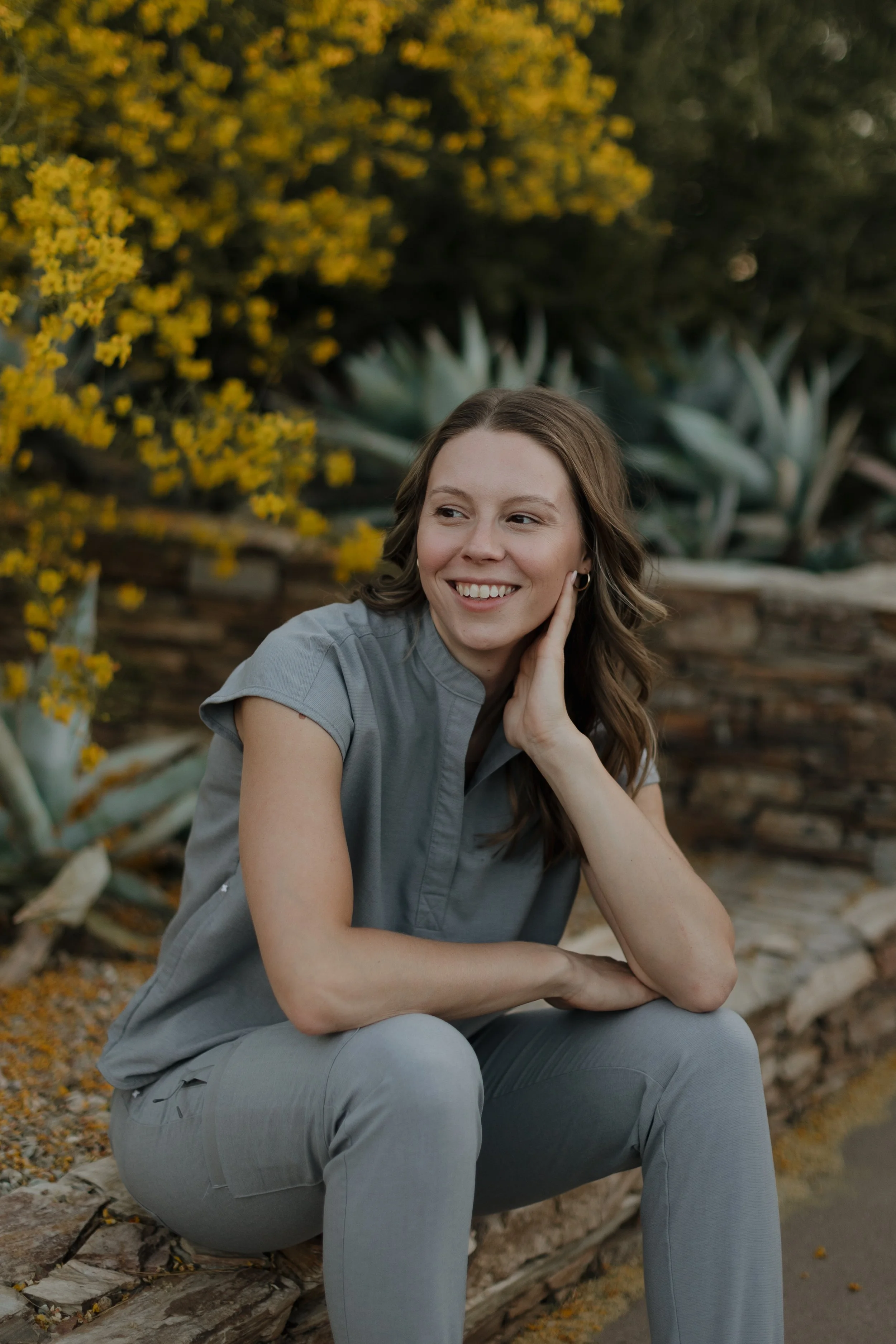 A female naturopathic doctor with brown hair and a light gray short-sleeve shirt, sitting outdoors with a smile, surrounded by yellow flowering bushes and agave plants in the background.
