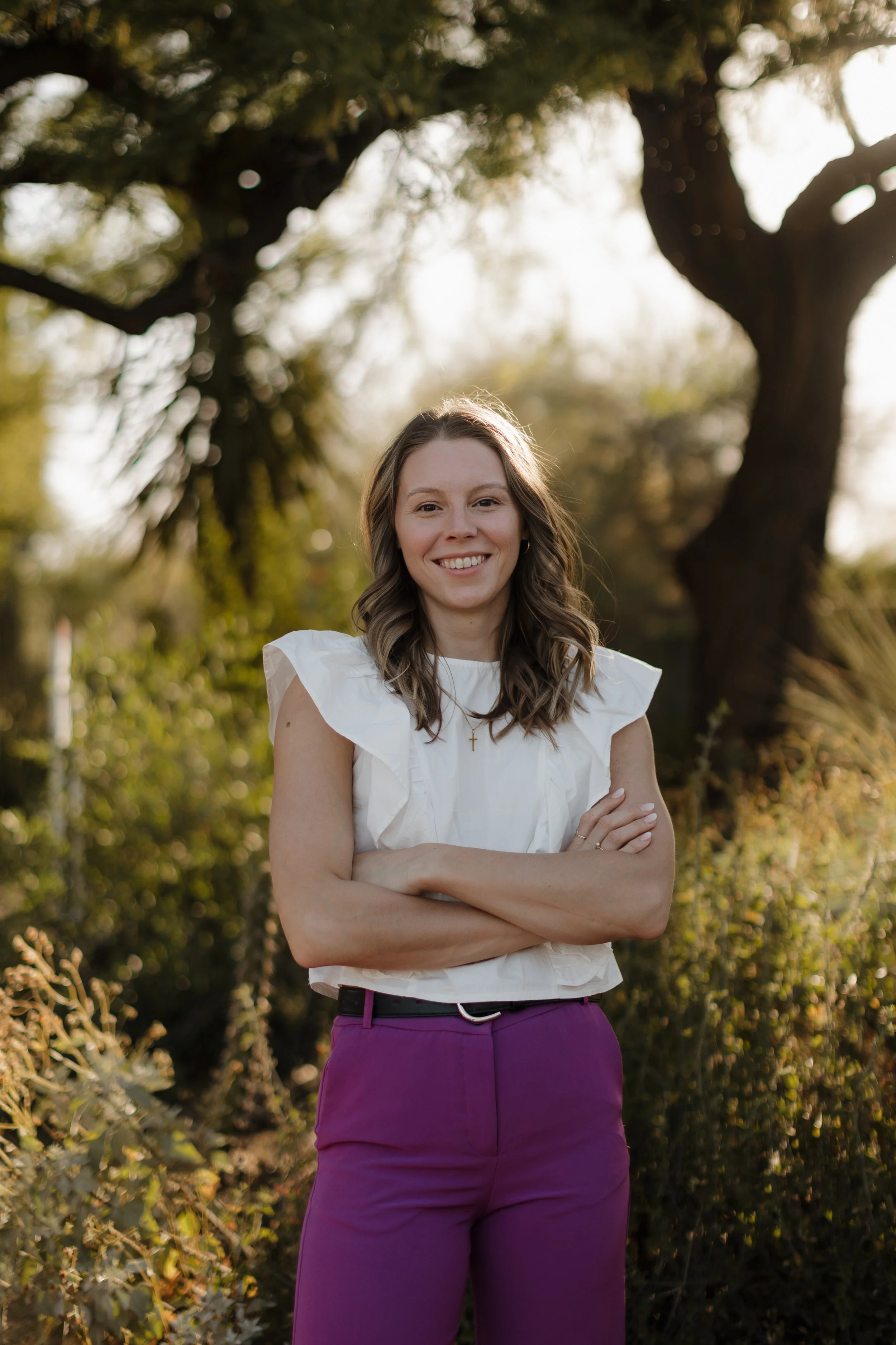 A female Naturopathic Doctor with light skin and wavy brown hair smiling and standing outdoors in front of a large tree.