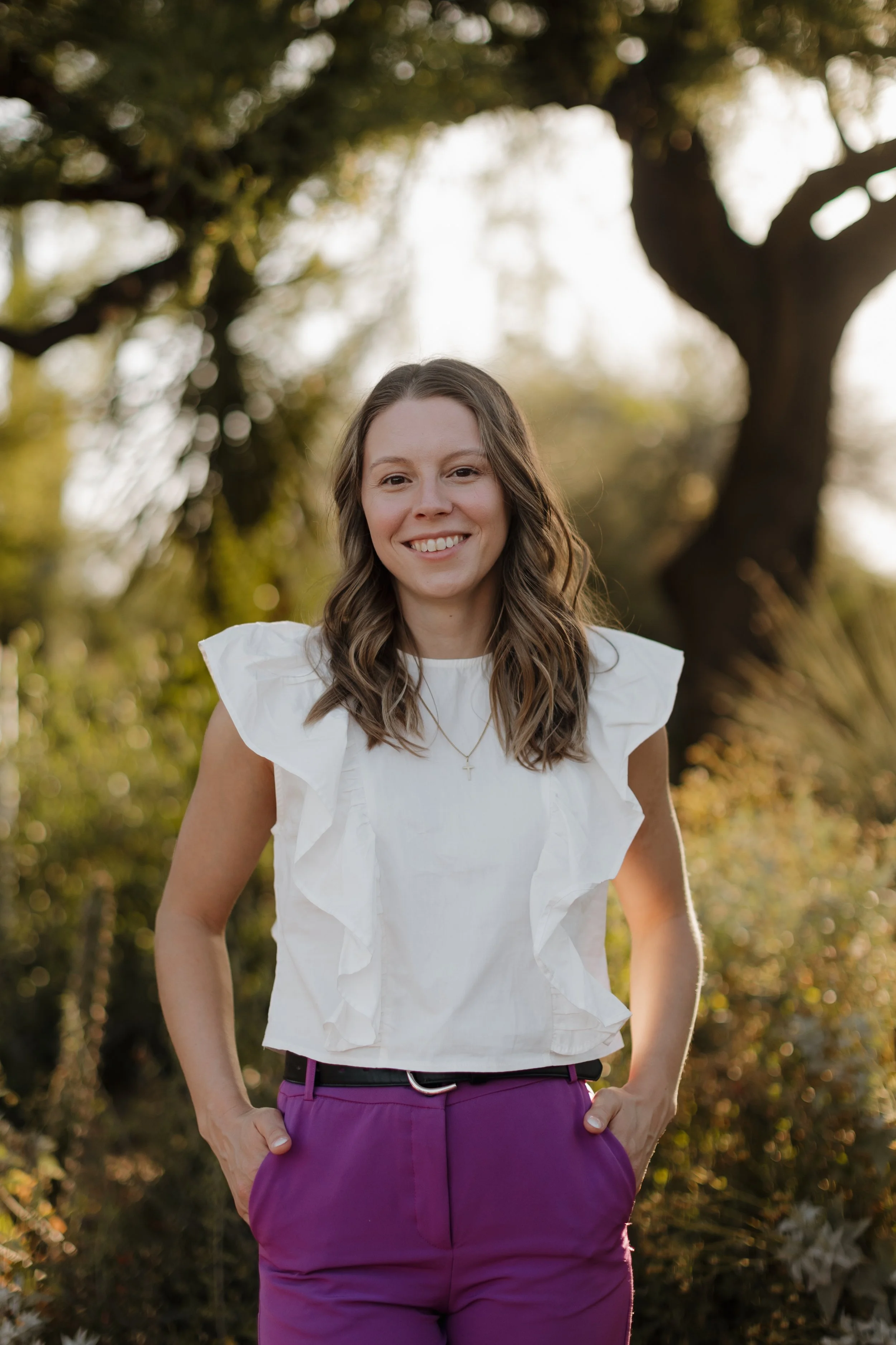A smiling female Naturopathic Doctor with wavy brown hair wearing a white ruffled top and purple pants standing outdoors with trees and sunlight in the background.