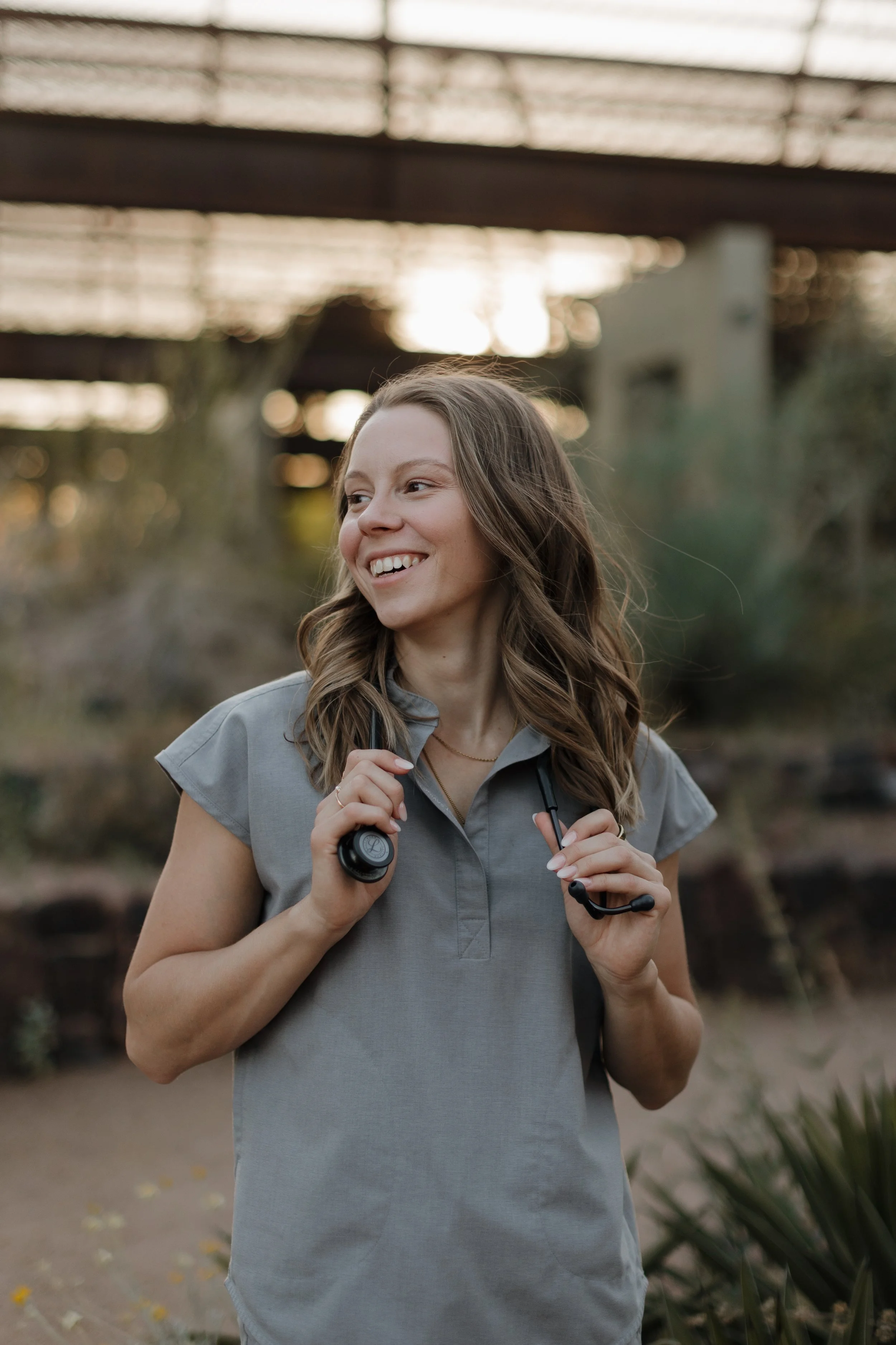 A female naturopathic doctor is holding a stethoscope around her neck while outdoors during sunset or sunrise, with a bridge or structure in the background.
