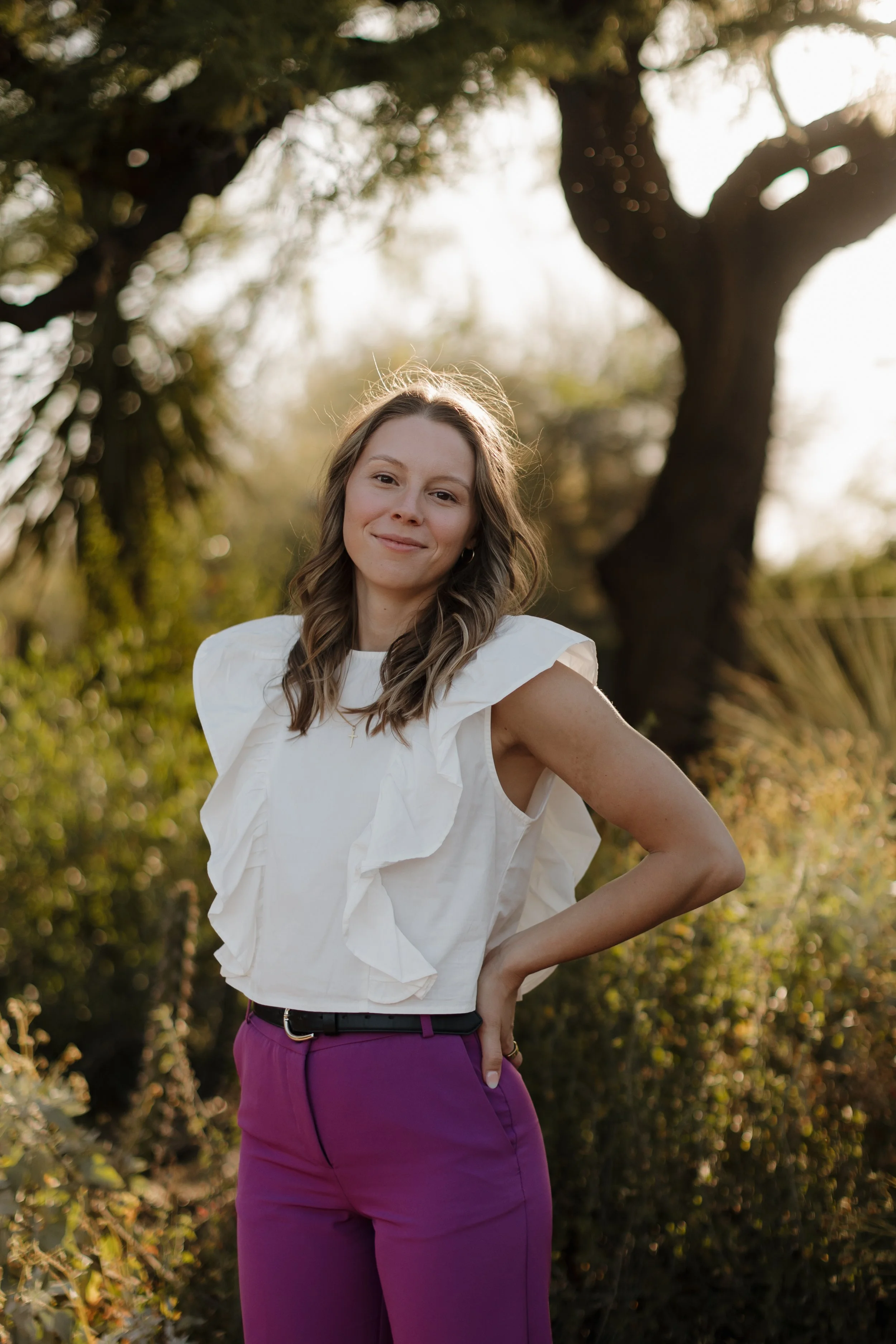 A female naturopathic doctor with light skin and wavy light brown hair standing outdoors in front of a large tree, smiling, wearing a white ruffled sleeveless top and purple pants, with sunlight filtering through the trees.