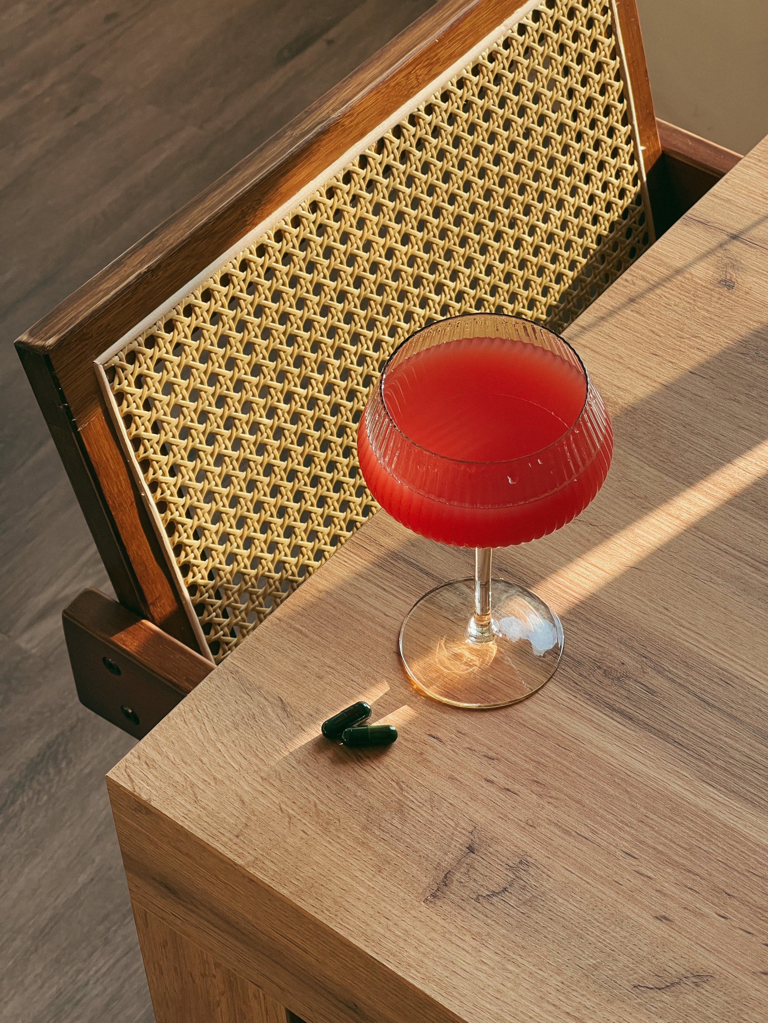 A glass of red cocktail on a wooden table, with a woven wooden chair back behind, and two green capsules on the table beside the glass for naturopathic doctor brand
