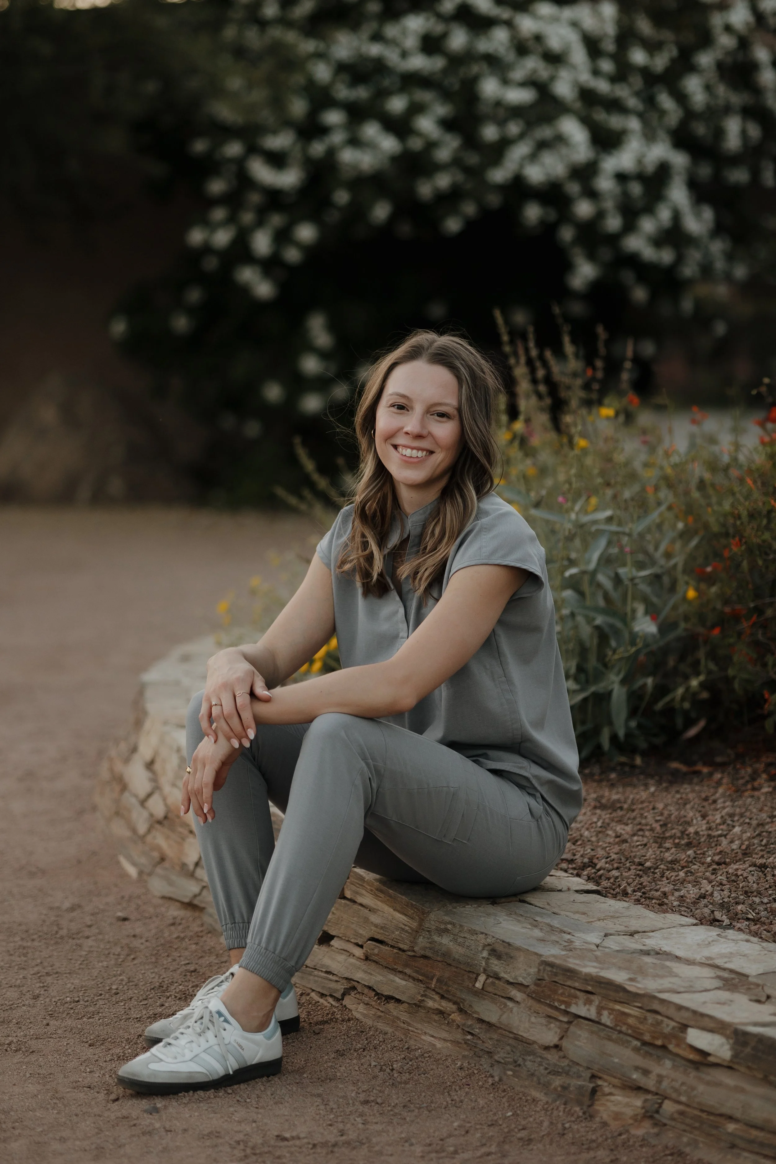 A female naturopathic doctor sitting on a log outdoors, smiling at the camera, with a background of black bushes with white flowers.