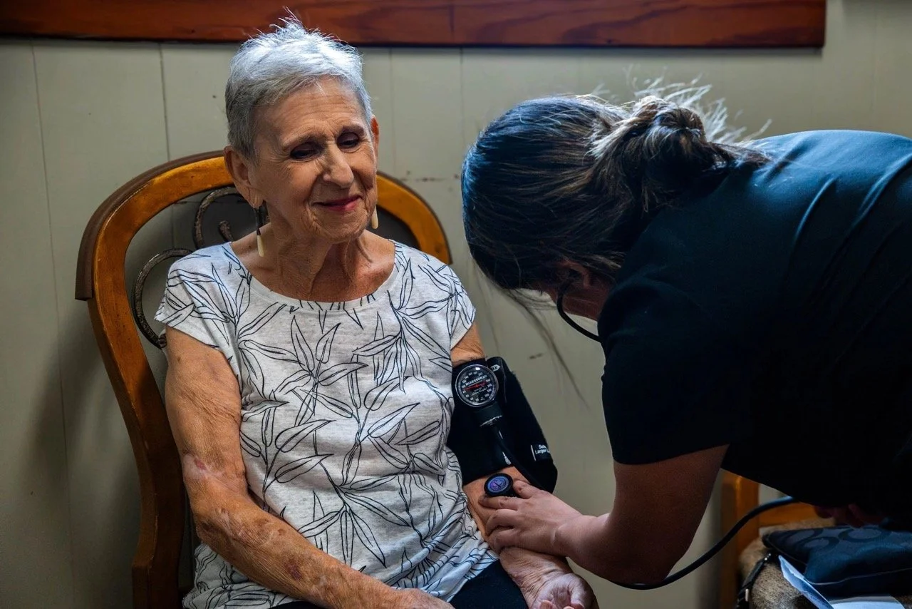 A healthcare worker using a blood pressure cuff on an elderly woman seated in a chair, in a room with beige walls and wooden paneling.