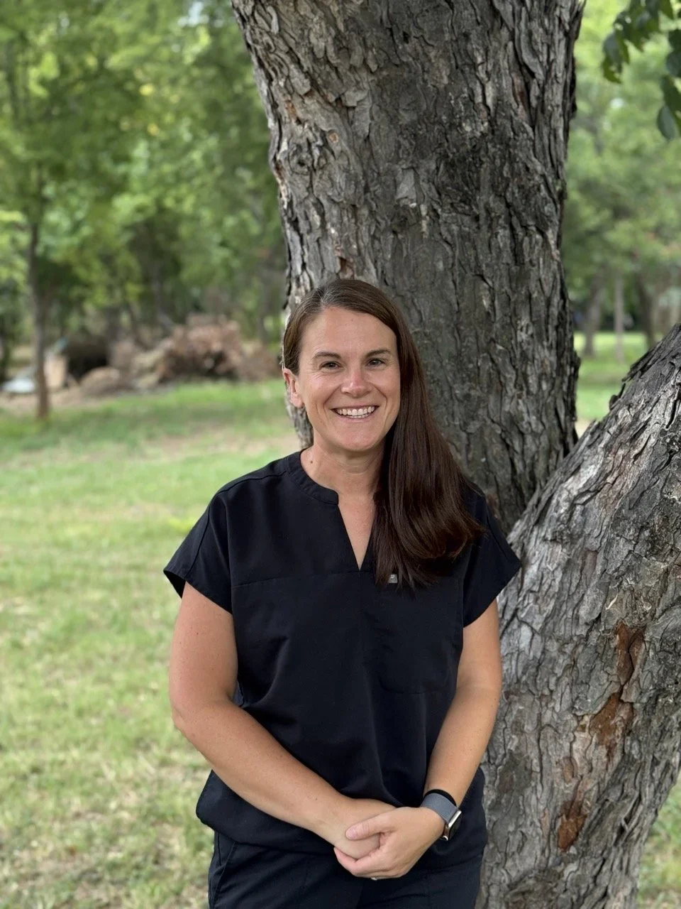 A woman with long brown hair smiling, standing outdoors next to a large tree with textured bark, in a park with green grass and trees in the background.