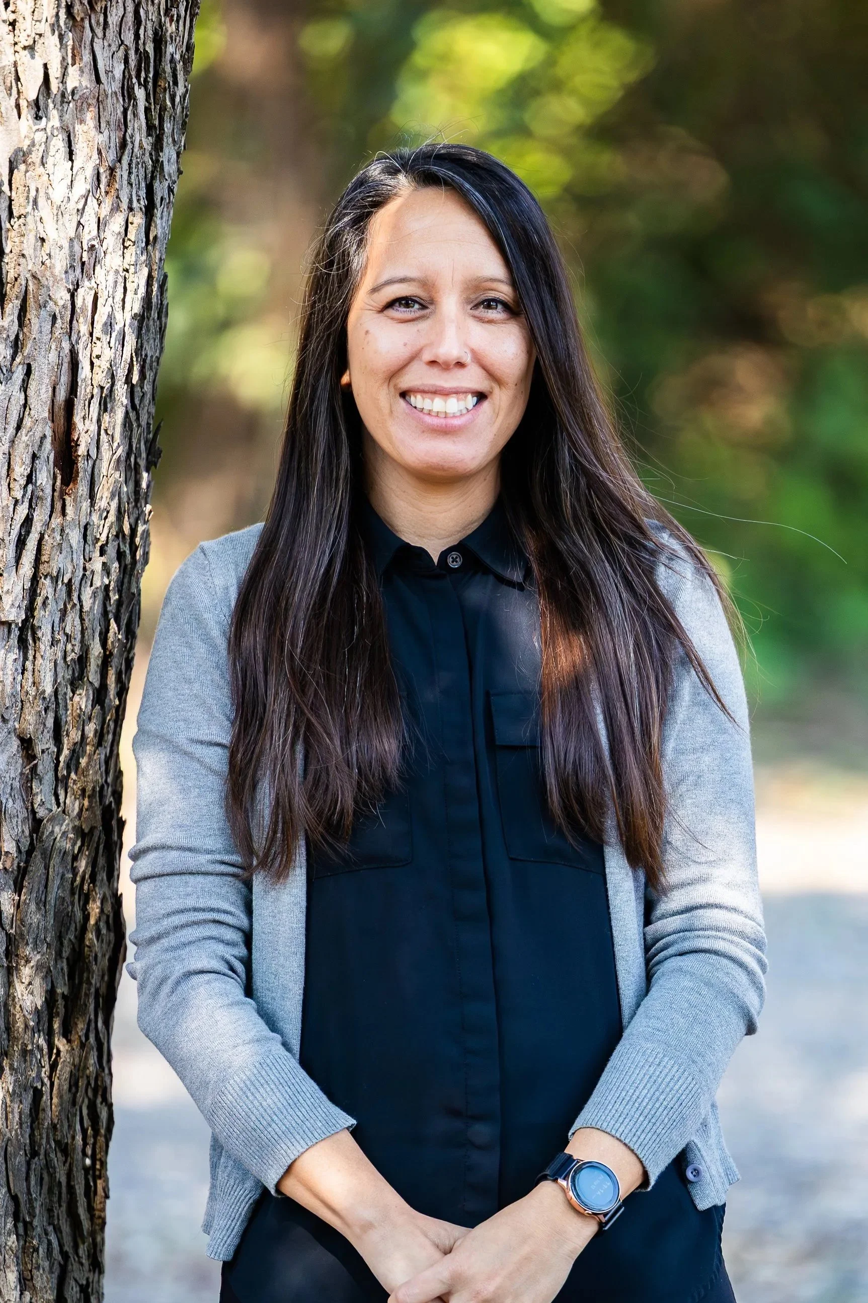 A woman with long dark hair smiling outdoors next to a tree with a blurred green background.
