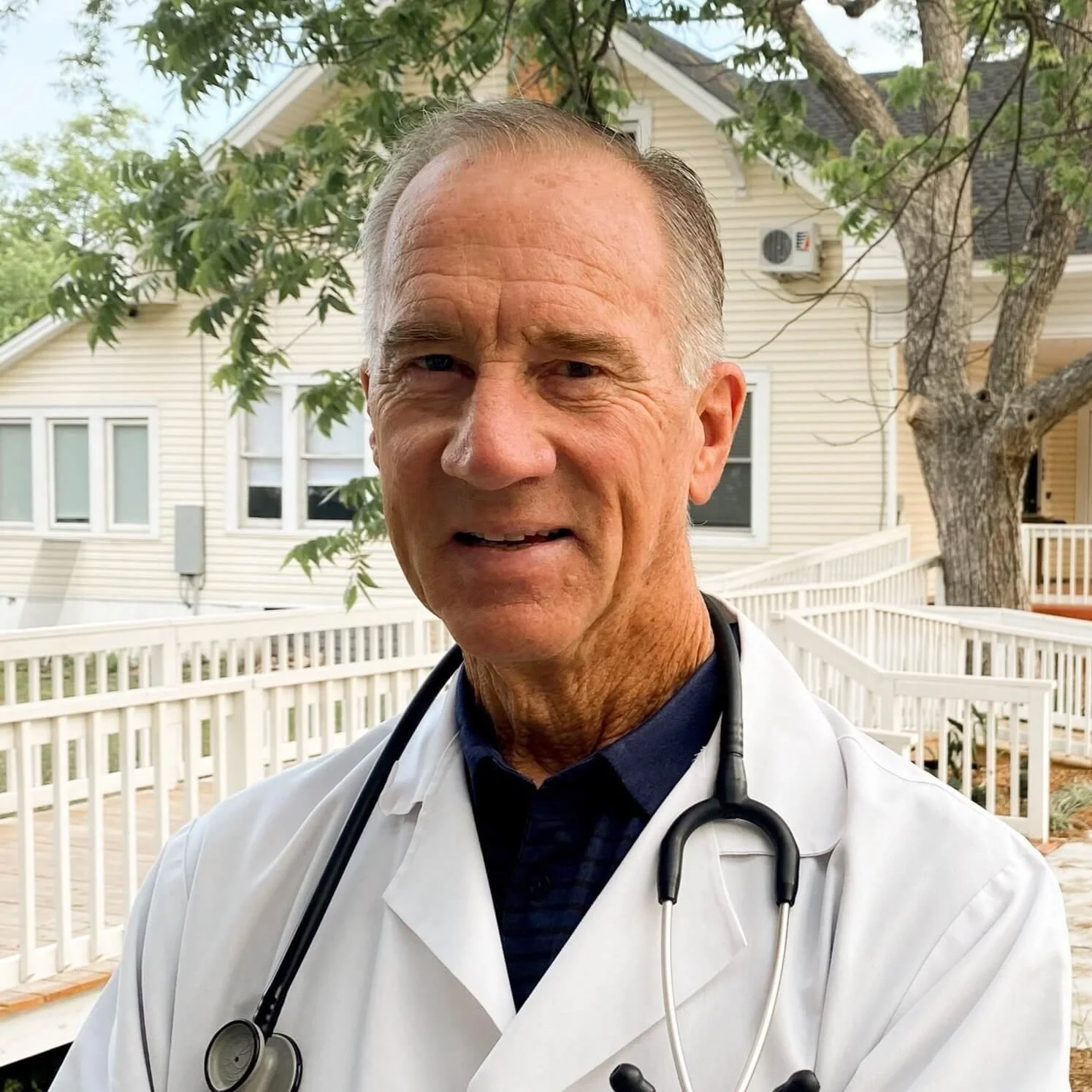 A male doctor wearing a white coat and stethoscope standing outdoors with a house and trees in the background.