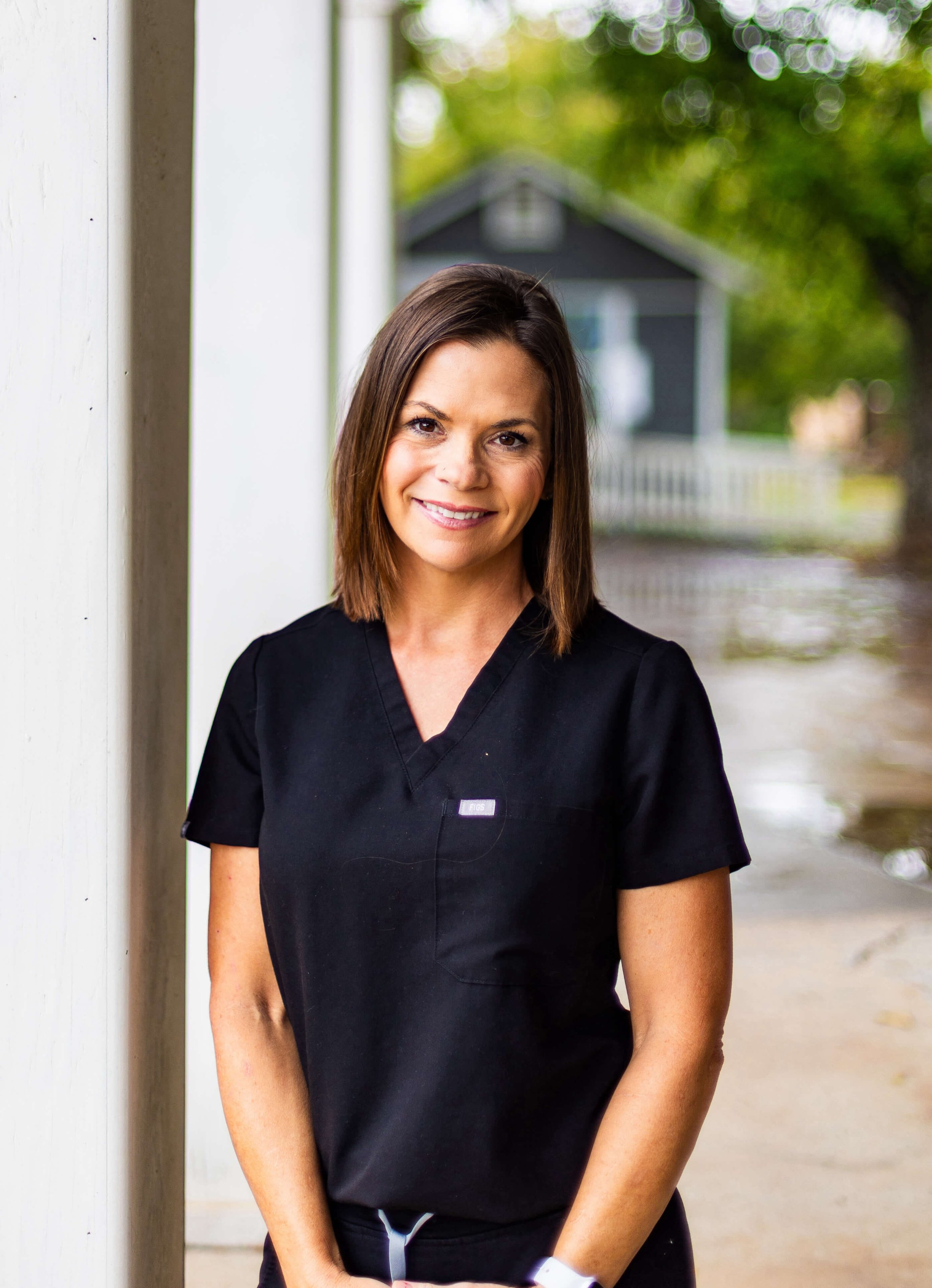 Smiling woman wearing black scrubs standing outside near a white post with a blurred rustic building and trees in the background.