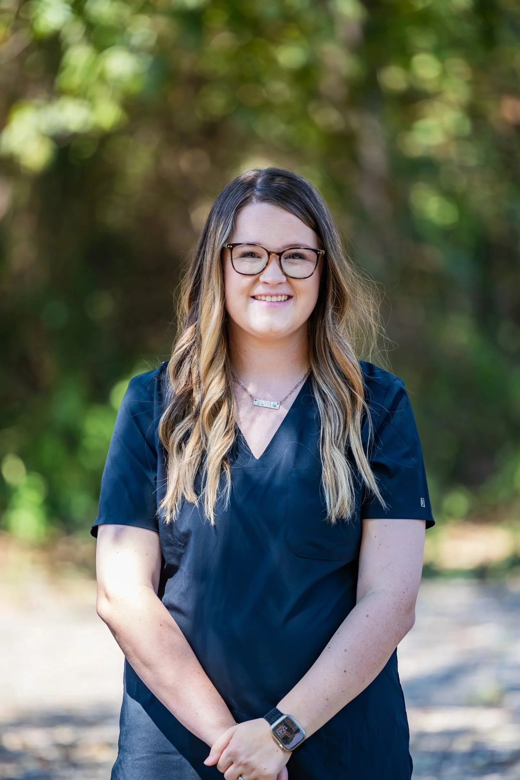 A woman with long, wavy hair, wearing glasses and a navy blue top, standing outdoors with blurred green trees in the background, smiling at the camera.