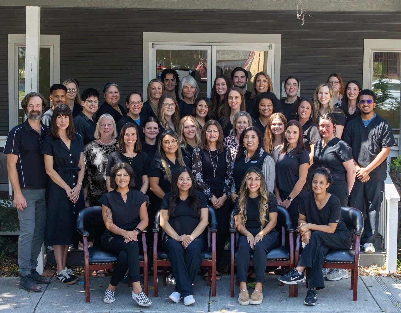Group photo of diverse people standing and sitting in front of a house, dressed in black and dark clothing, smiling for the camera.
