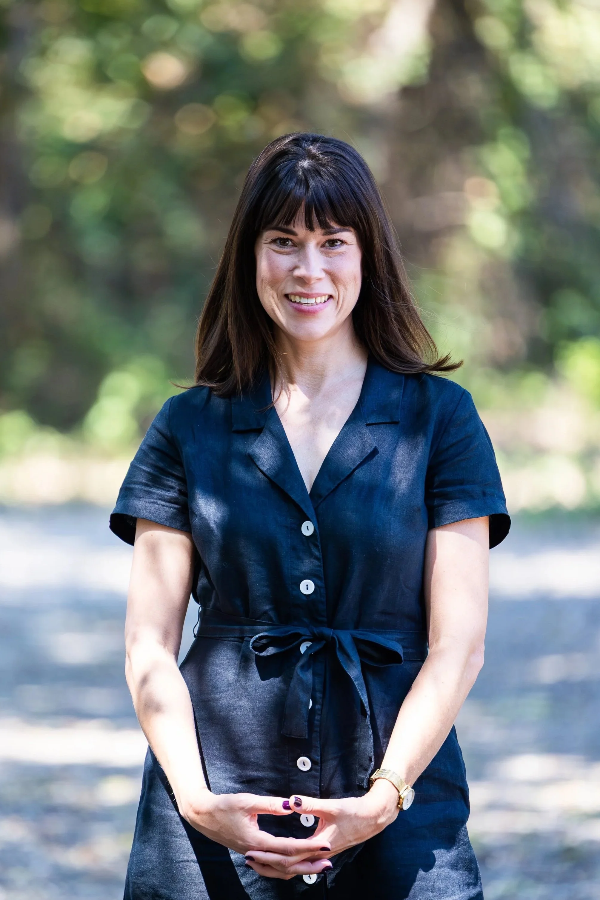 A woman with shoulder-length dark hair and bangs, smiling outdoors, wearing a navy blue button-up dress with a tied waist, in a natural setting with blurred green foliage background.