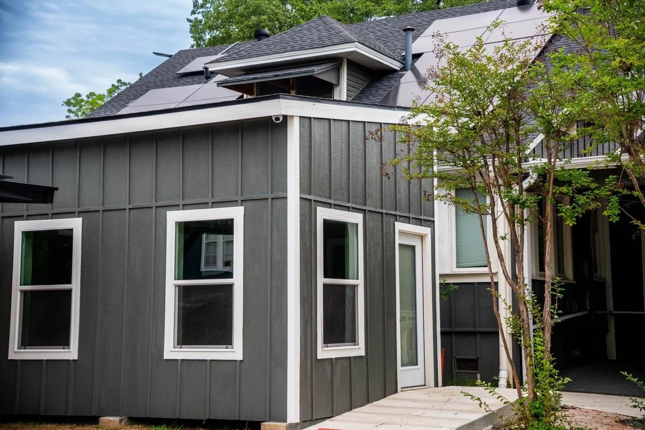 Gray modern house with white window frames, a small door, a tree on the right, and solar panels on the roof.