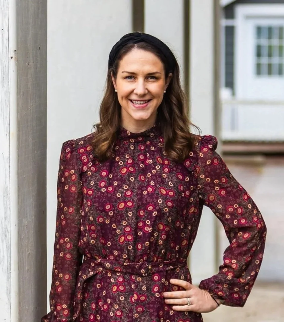 A woman with long brown hair, wearing a black headband and pearl earrings, stands outdoors next to a wooden post, smiling at the camera. She is dressed in a long-sleeved, patterned red and purple dress with small circular designs.