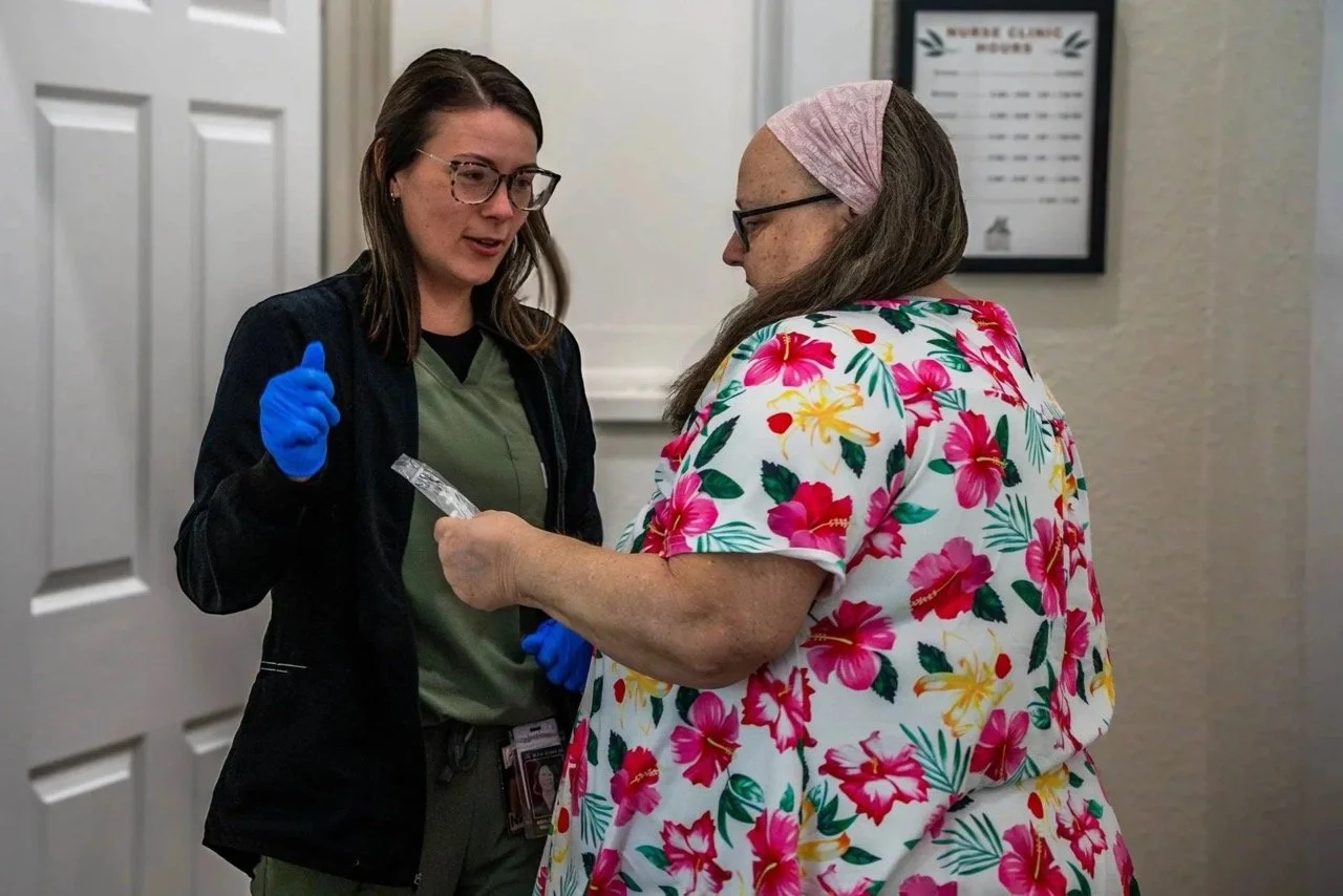A healthcare worker, wearing glasses, a black jacket, and blue gloves, is handing a package or medication to an elderly woman with long gray hair, glasses, and a pink headscarf, who is dressed in a colorful Hawaiian style shirt. They are standing in a room with a white door and a framed health clinic sign on the wall.