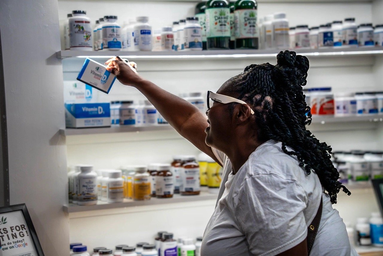 A woman with braids and glasses reaching for a bottle of Vitamin D supplement on a store shelf filled with vitamins and supplements.