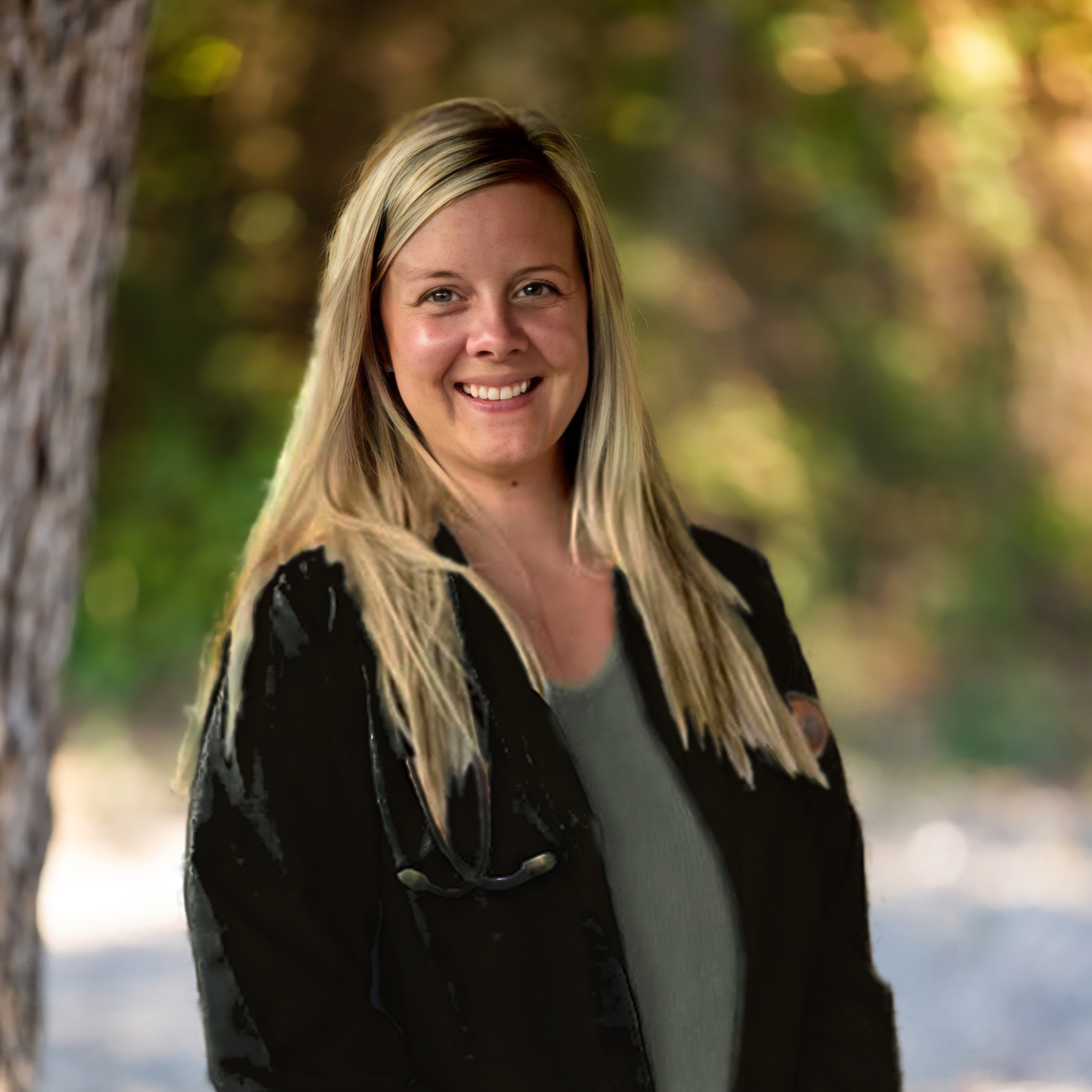 Close-up photo of a smiling woman with blond hair outdoors, with blurred trees and sunlight in the background.