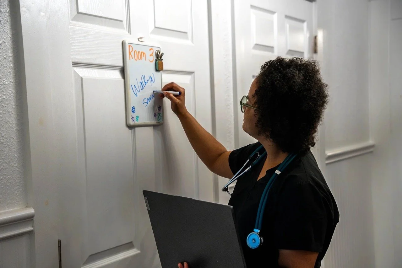 A female healthcare professional with curly hair, glasses, and a stethoscope around her neck writes on a whiteboard attached to a white door in a hallway. The whiteboard has