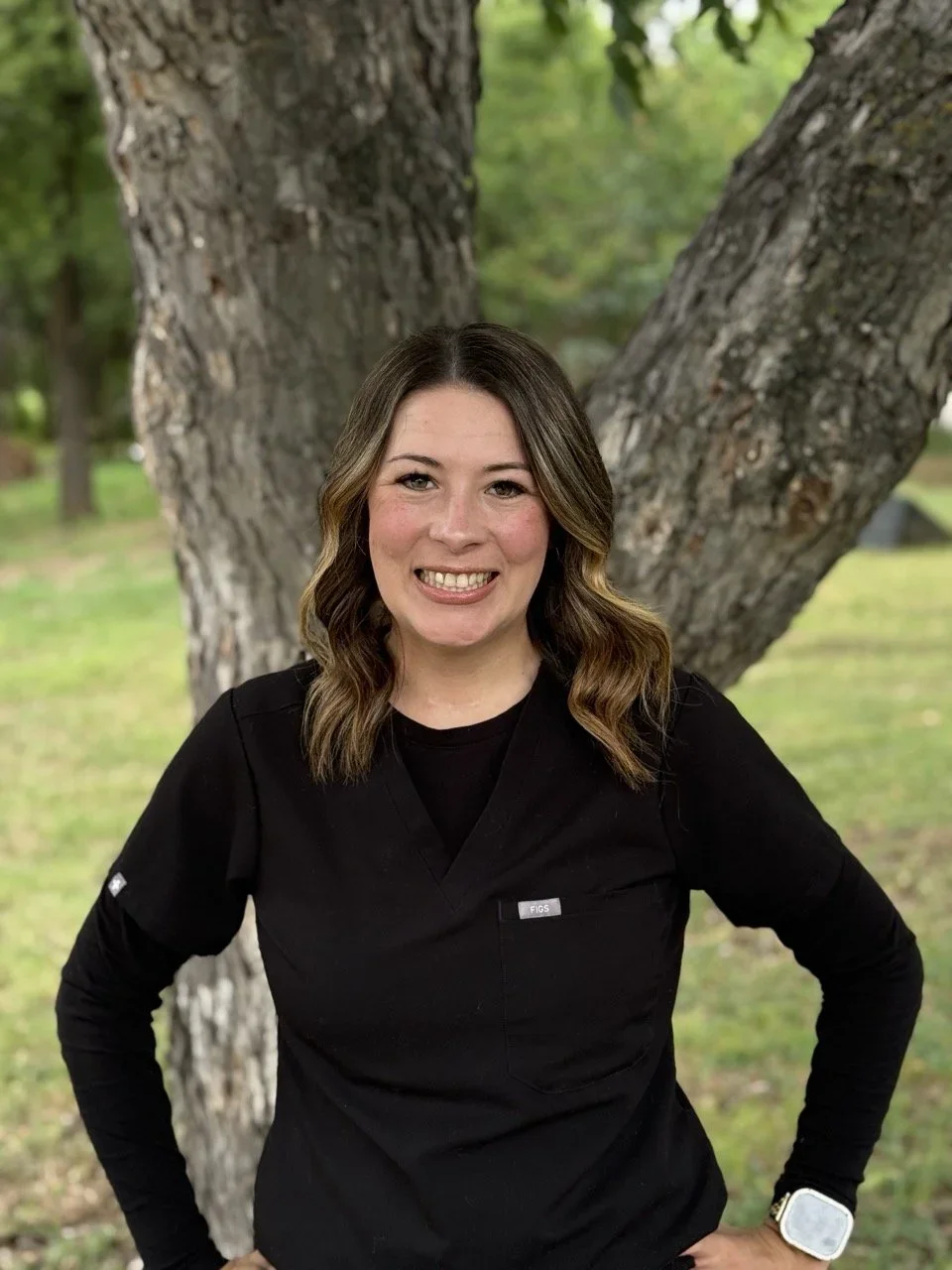 A woman in black scrubs with a name badge, standing outdoors with trees in the background, smiling at the camera.