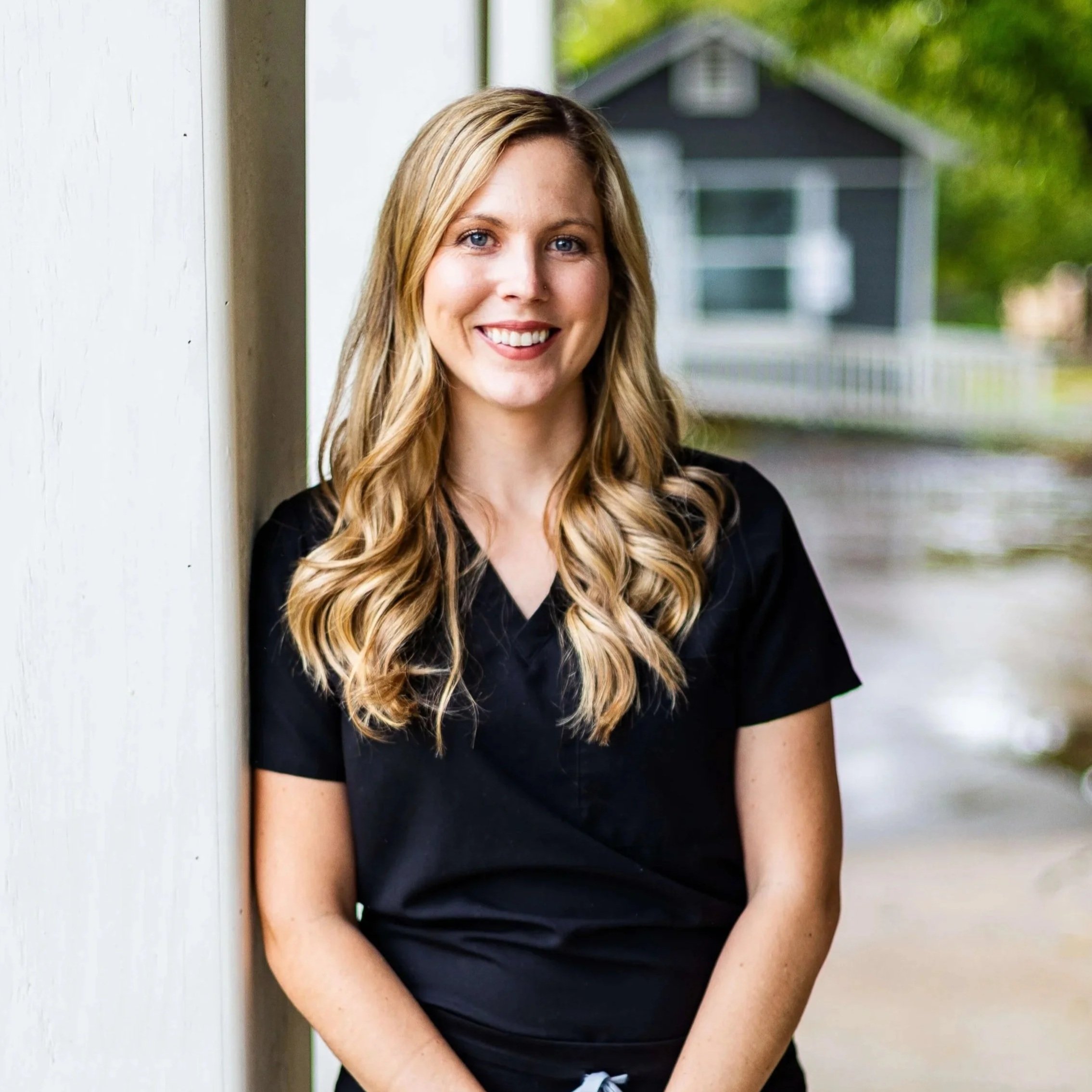 A woman with long wavy blonde hair, wearing a black shirt, is smiling and standing outdoors near a white wall with a background of green trees and a small gray house.