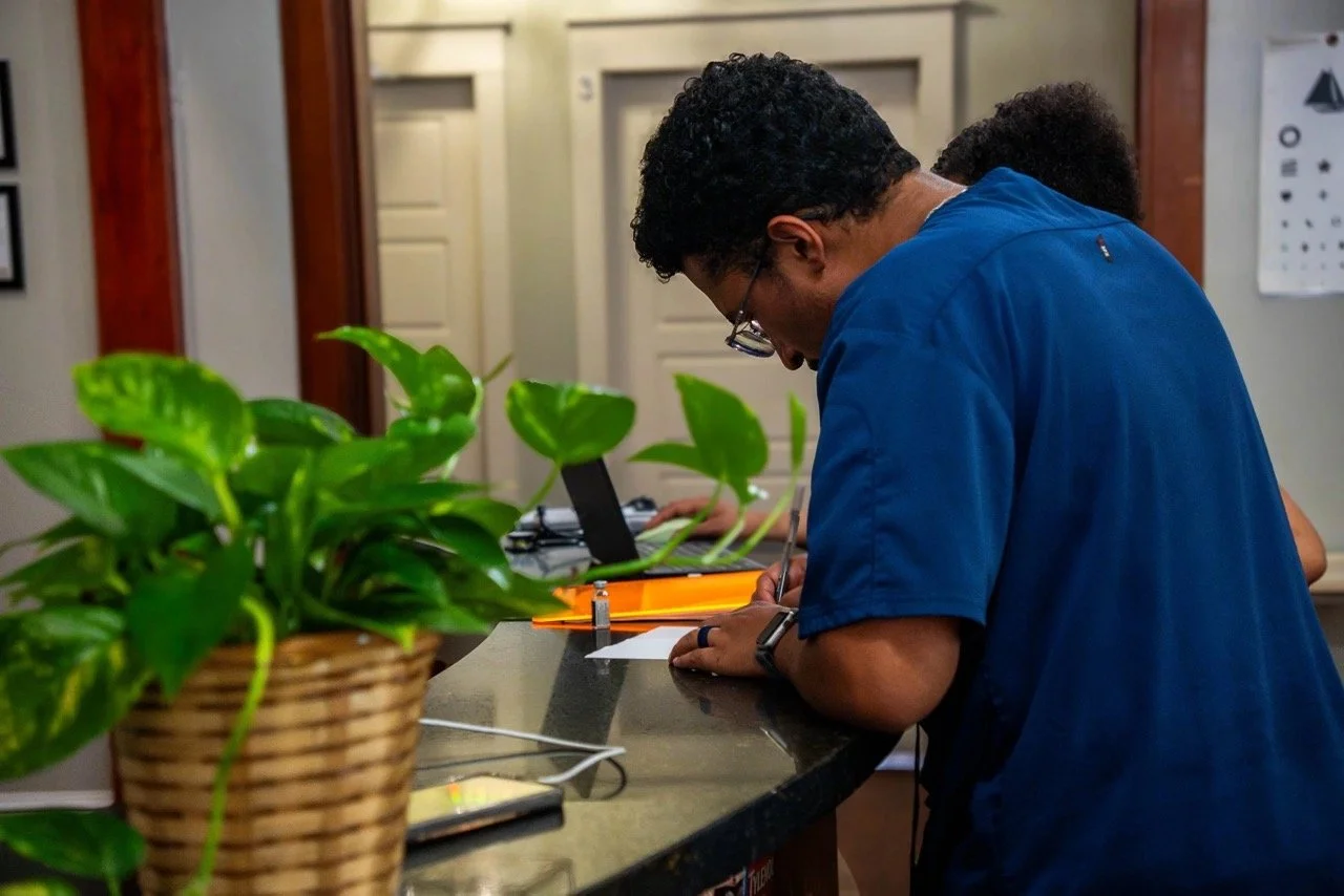 A man in a blue uniform writing on a piece of paper at a counter with green plants near him.