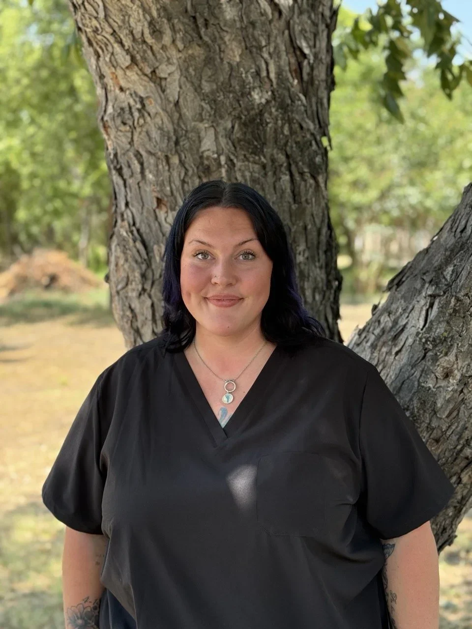 A woman with black hair, wearing a black top and necklace, standing outdoors in front of a large tree trunk with greenery in the background.
