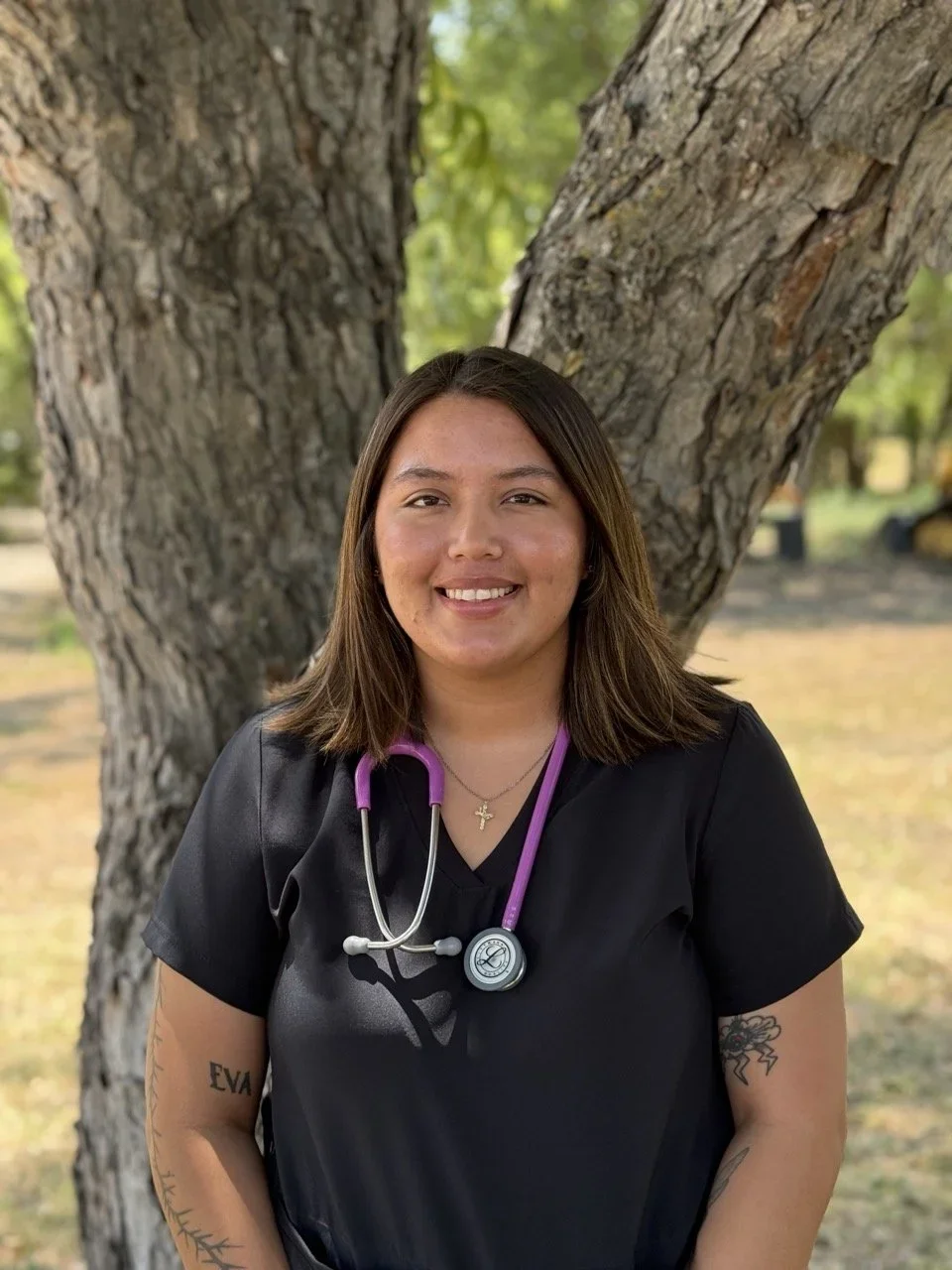 A woman in a black medical uniform wearing a purple stethoscope around her neck, standing outdoors in front of a large tree with green leaves in the background, smiling at the camera.